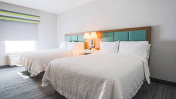 Een hotelkamer met twee bedden, een hoofdeinde met een blauwgroen kussen, een lamp en een raam met zonwering.