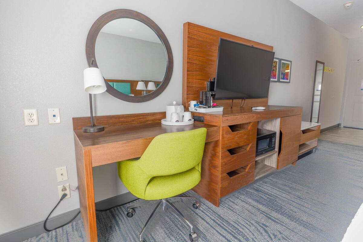A hotel room with a wooden desk, a green chair, a flat-screen TV, a mirror, and a microwave.