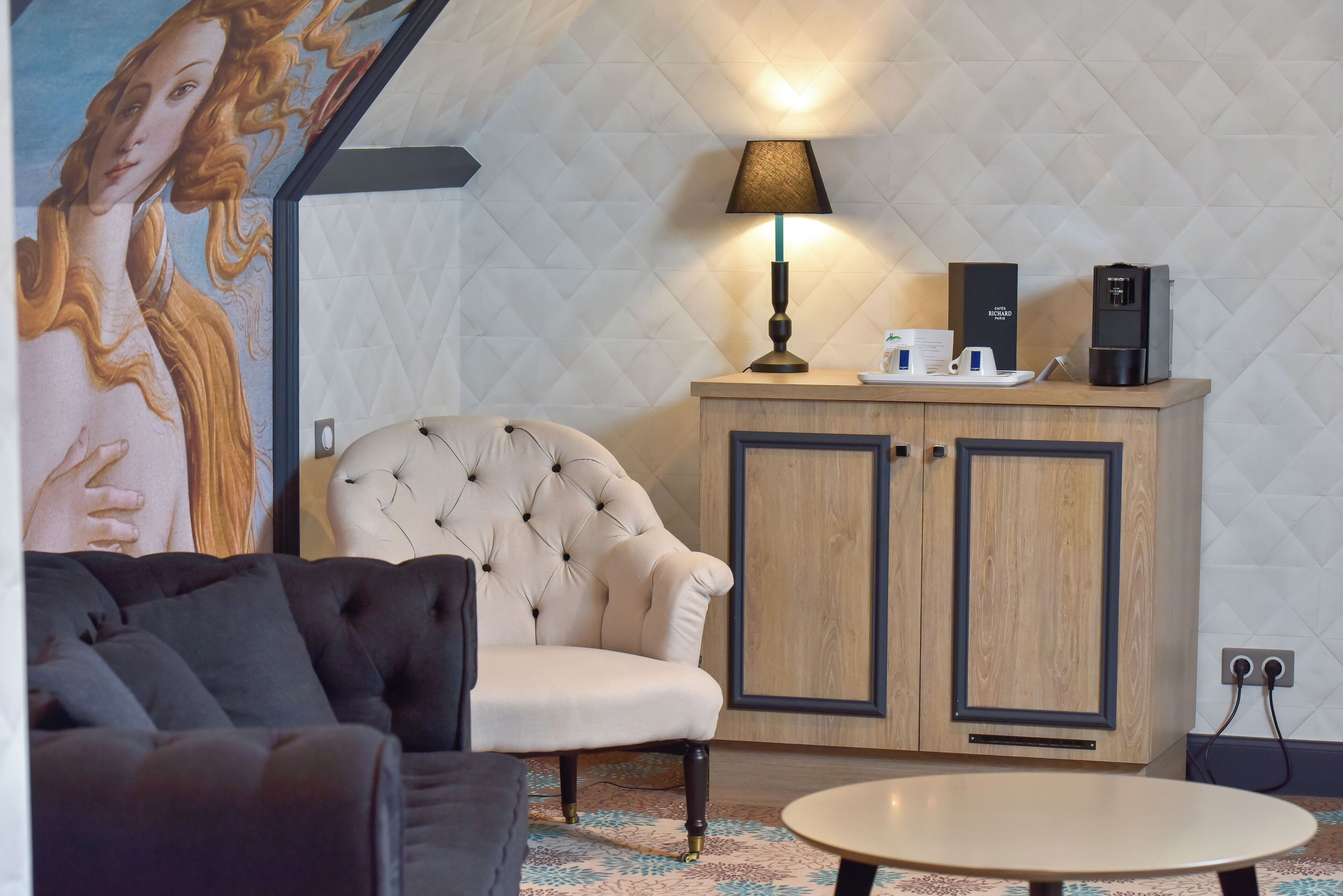 A modern room with a tufted armchair, a round table, a cabinet with a coffee machine, and a wall-mounted lamp.
