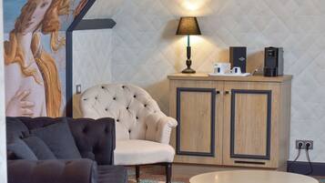A modern room with a tufted armchair, a round table, a cabinet with a coffee machine, and a wall-mounted lamp.