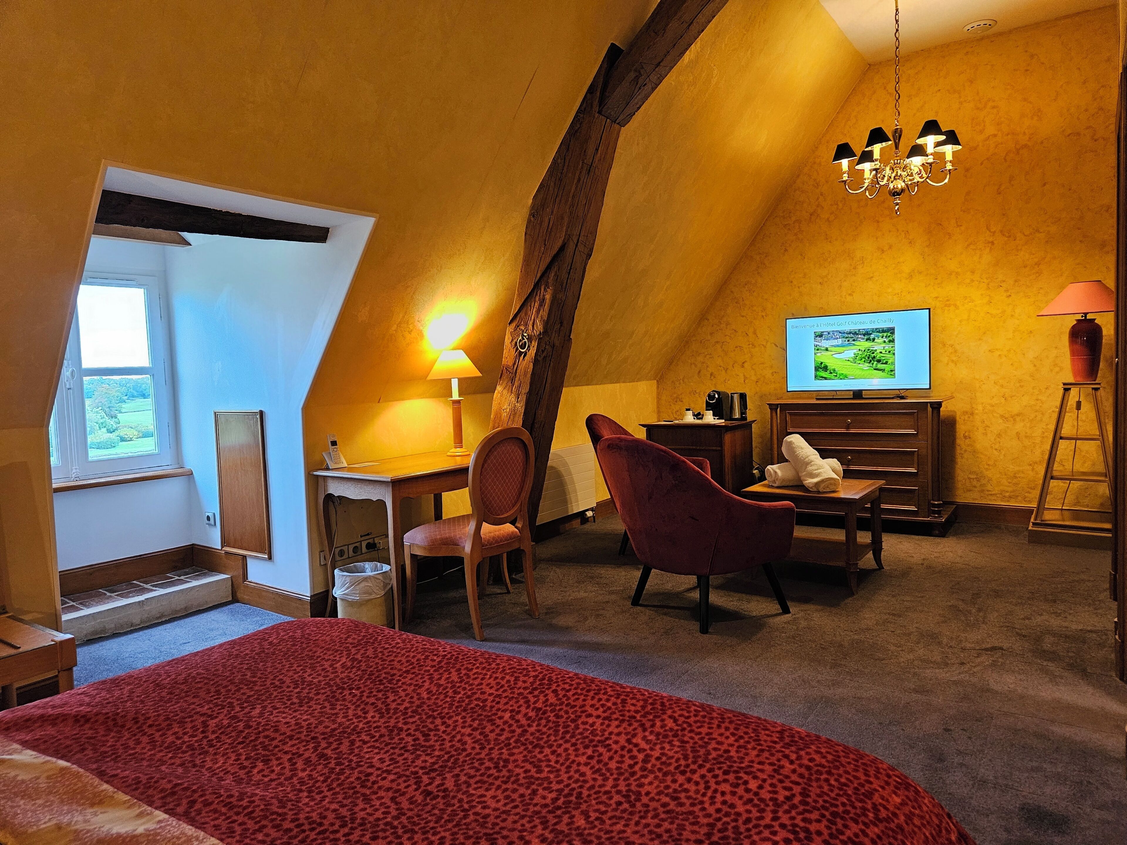 A cozy room with a bed, a desk, a chair, a television, and a chandelier.