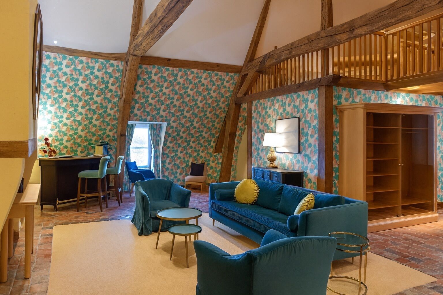A room with a high ceiling, exposed wooden beams, a patterned wallpaper, a blue sofa, a small round table, and a wooden cabinet.