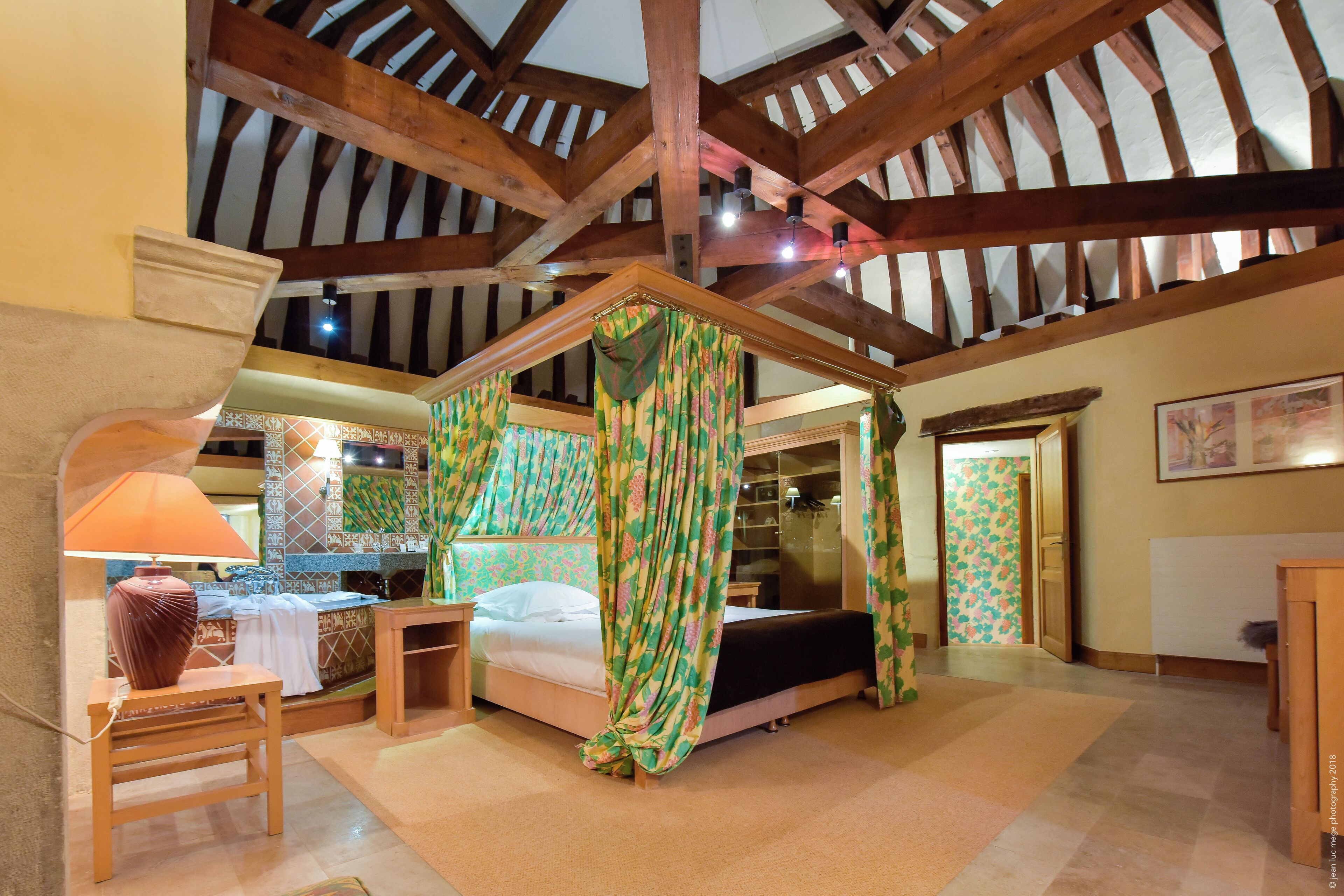 A room with a four-poster bed, a desk with a lamp, and a view of the upper floor.