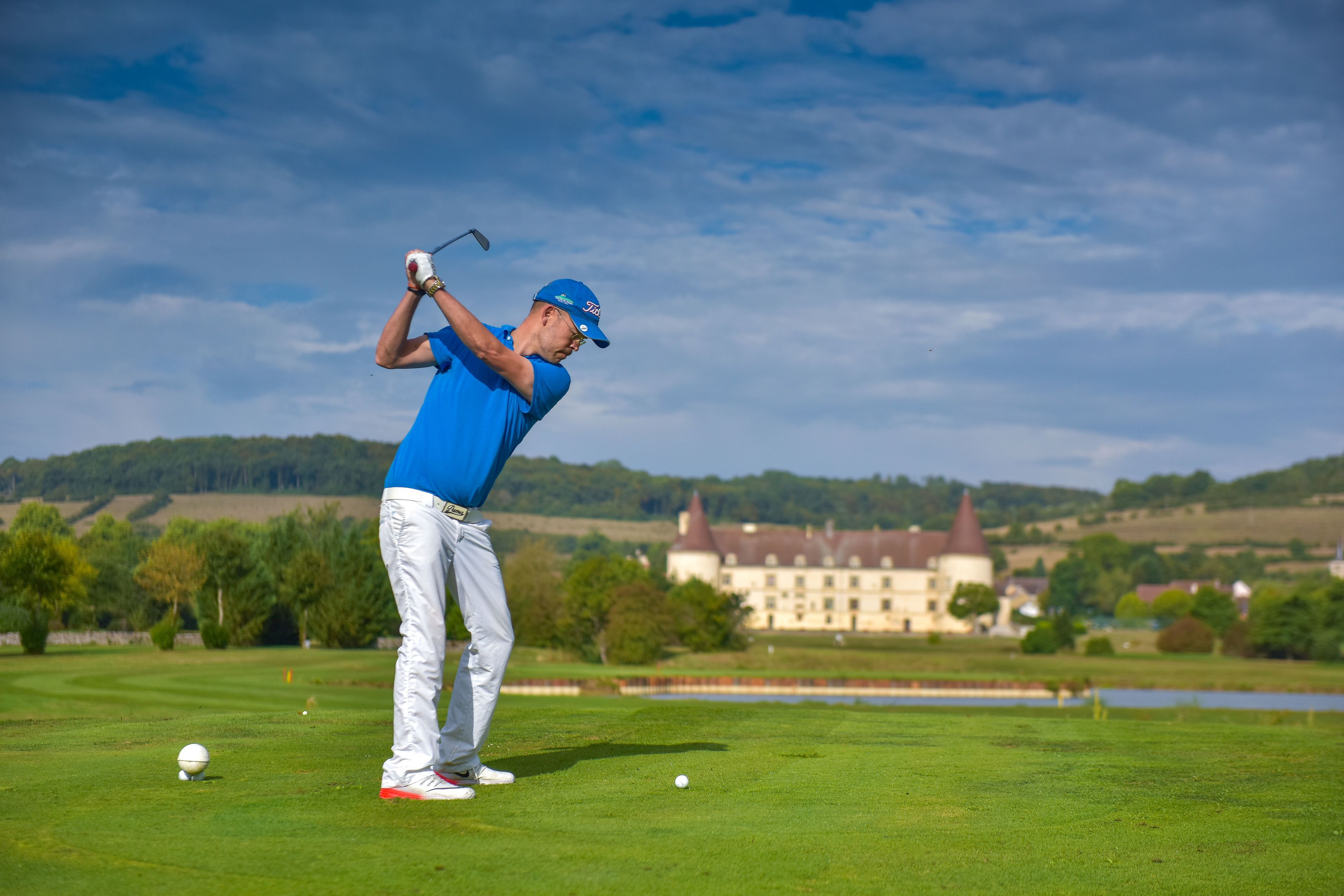 A golf course with a player in blue attire preparing to swing, a golf ball on the ground, and a building in the background.