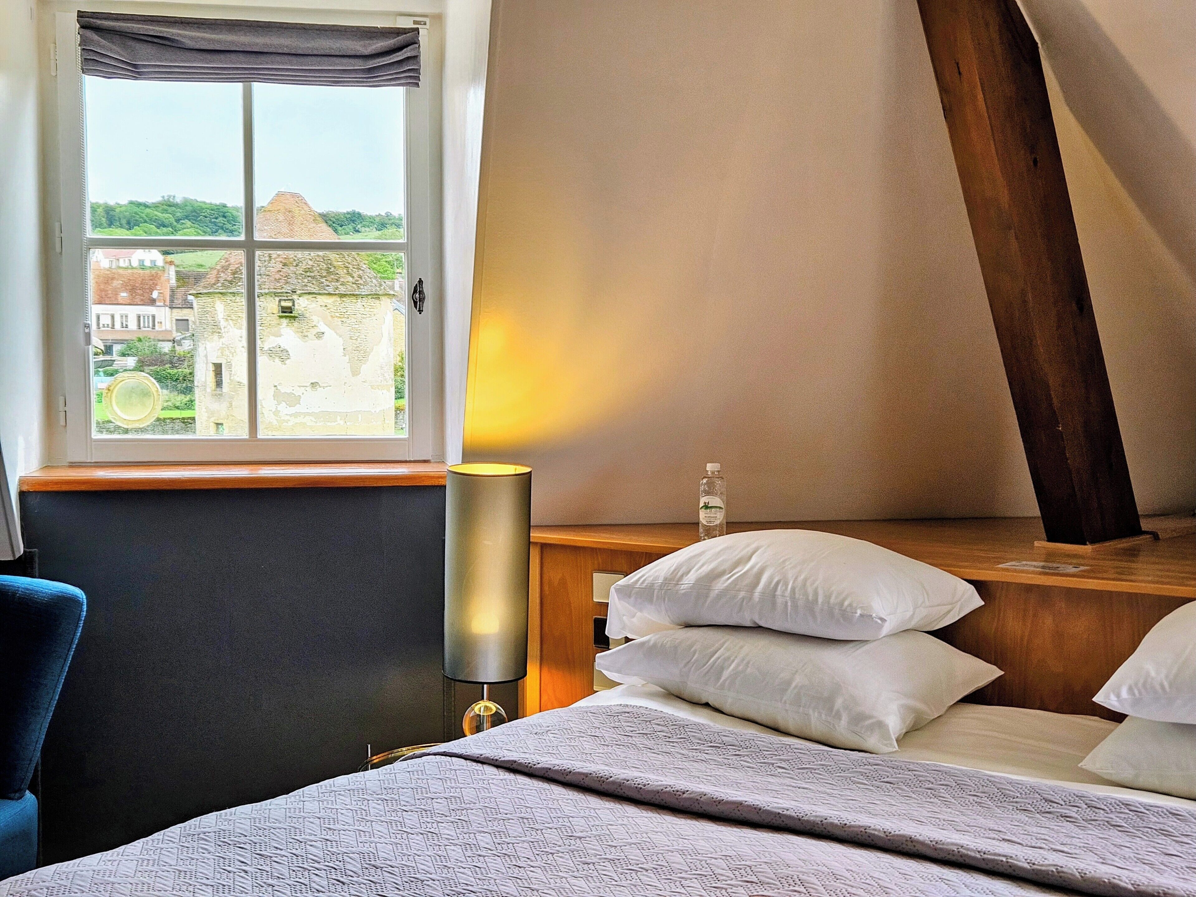 Ein Hotelzimmer mit Bett, Nachttisch mit Lampe, Fenster mit Blick auf Gebäude und einem Holzkopfteil.