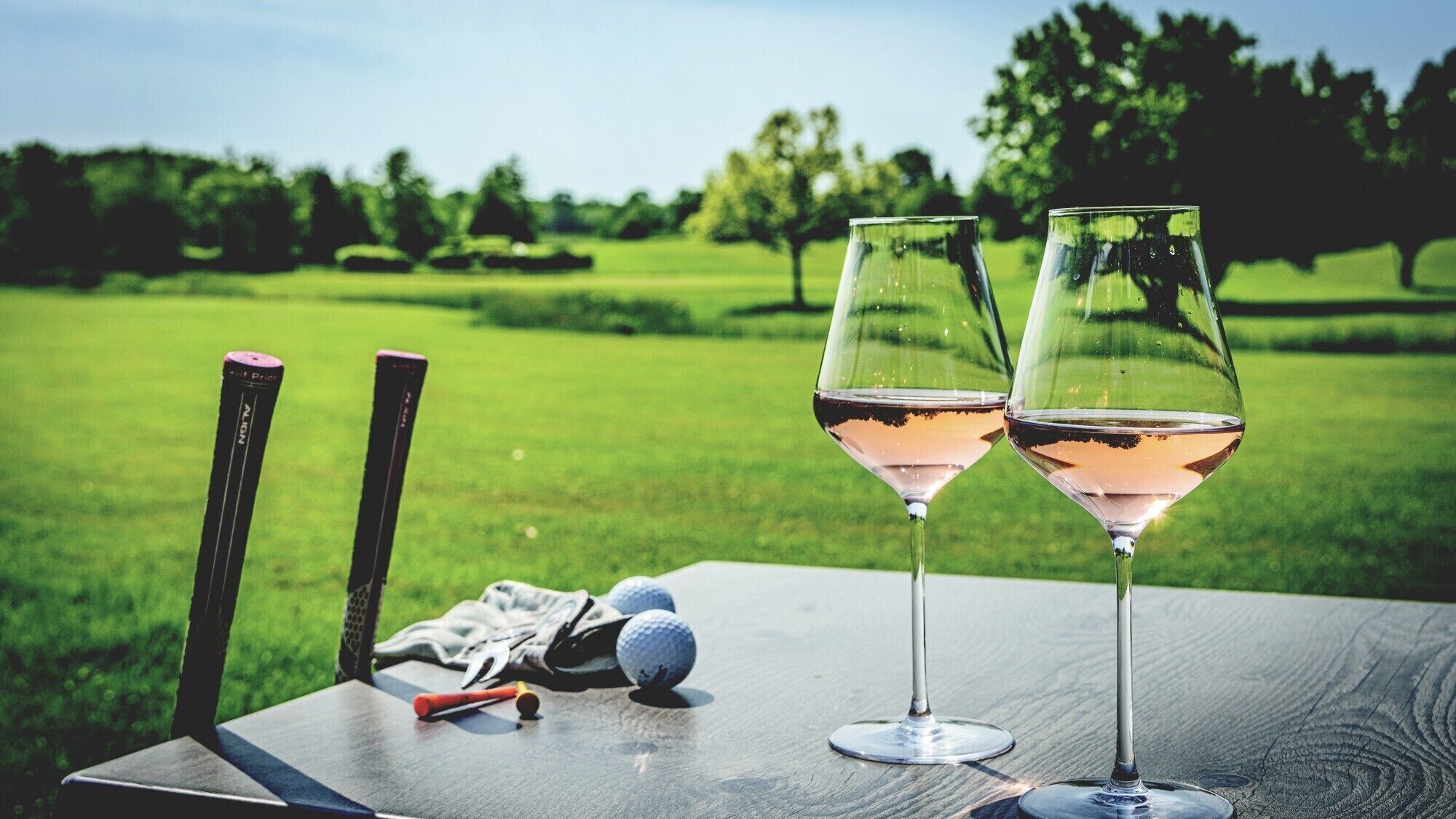 Two wine glasses on a table with golf equipment, set against a grassy field.