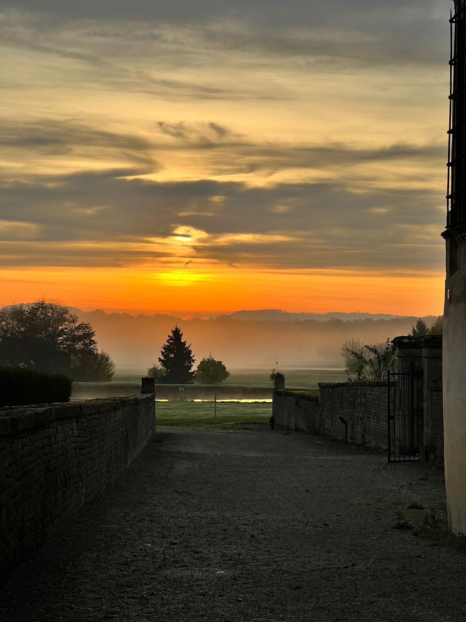 Un chemin de gravier mène à un bâtiment d’où l’on peut admirer un coucher de soleil sur un paysage brumeux.