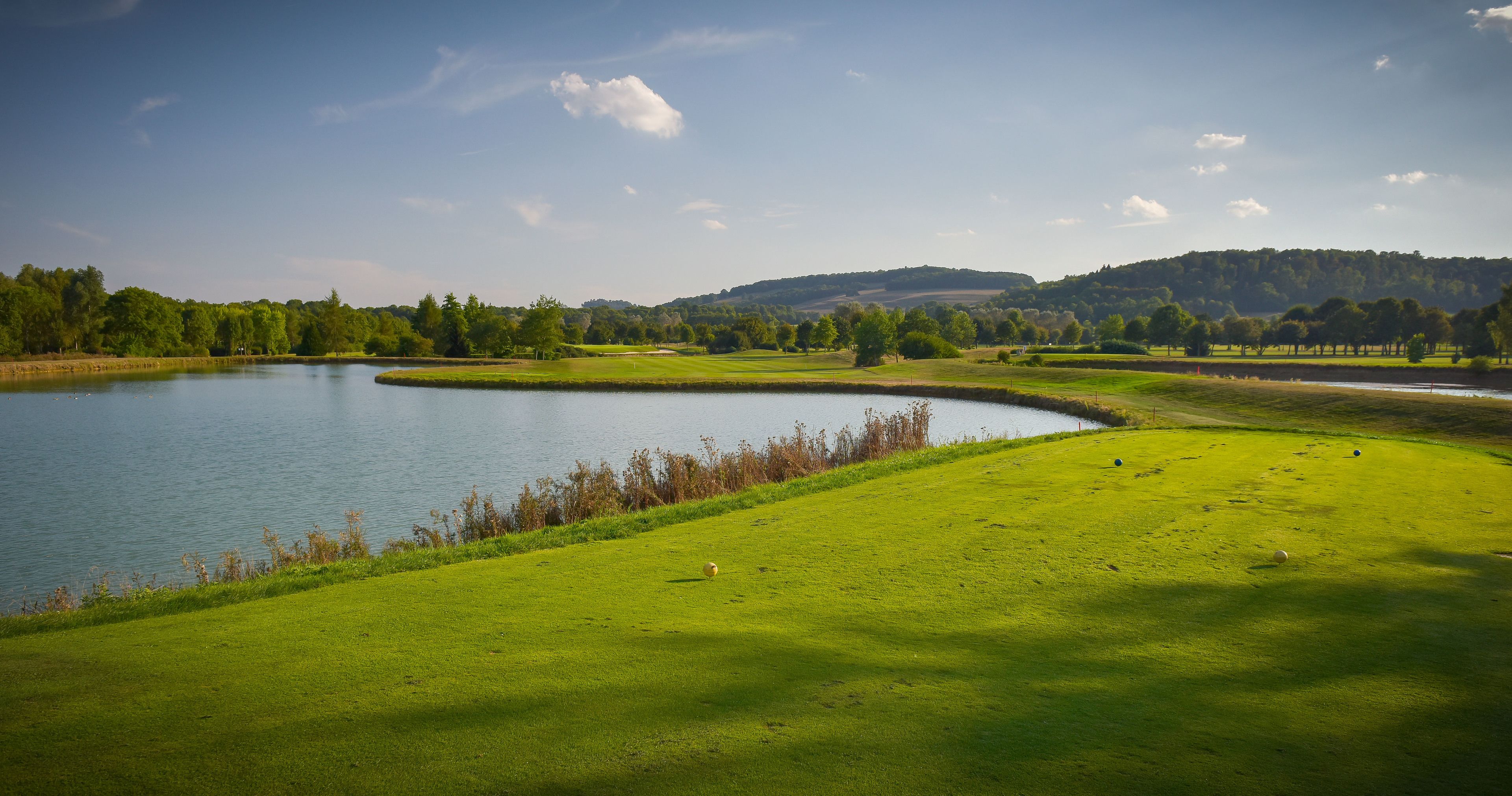 A golf course with a lake, surrounded by trees and hills.