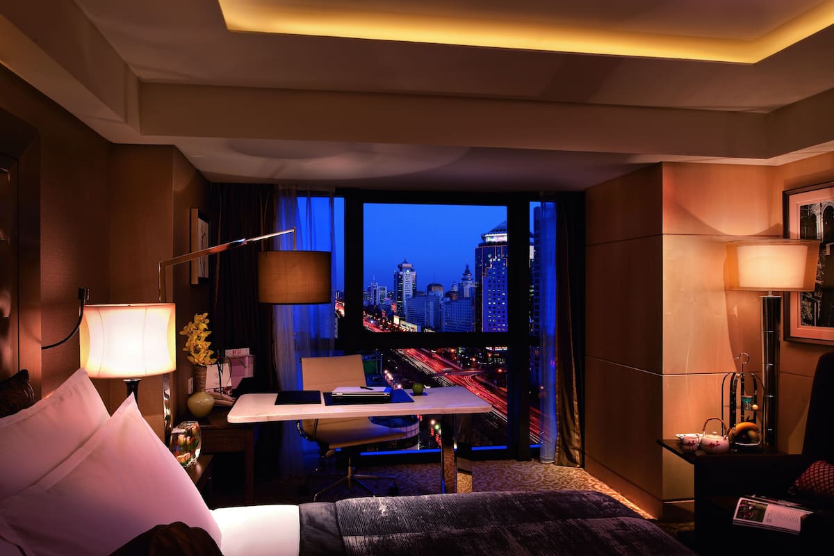 A hotel room with a city view, a desk, a chair, a bed, and a lamp.