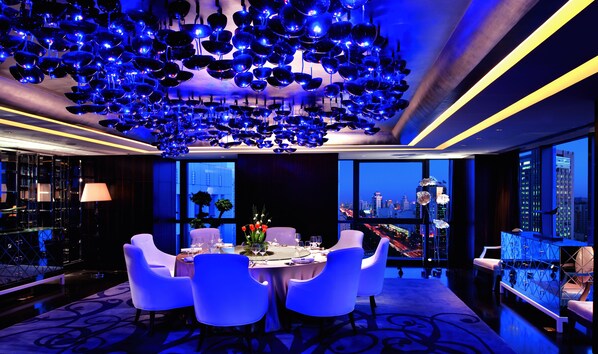 A modern dining area with a city view, featuring a table set for four, white chairs, and a unique ceiling light fixture.