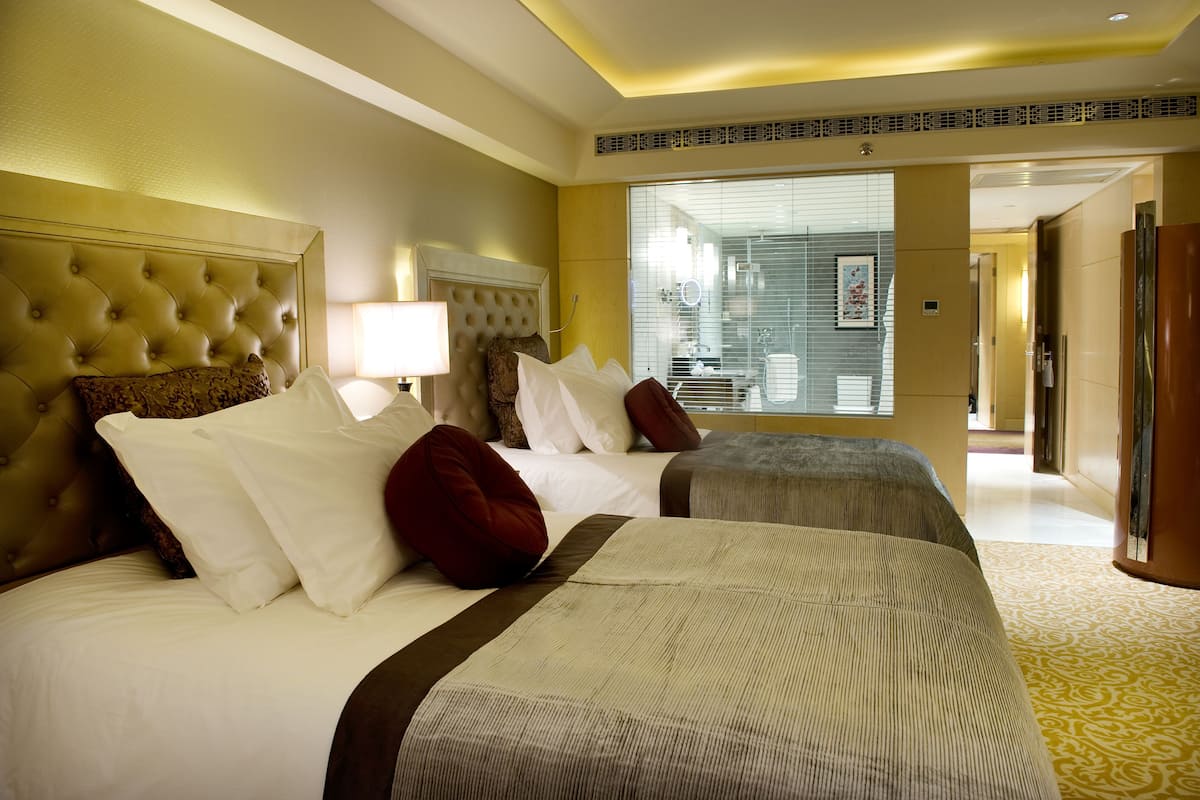A hotel room with two beds, a large headboard, a bedside table with a lamp, and a view of the bathroom through the window.