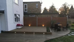A wooden deck with a table and chairs, surrounded by a white building and a brick wall. - The Firs Lodge (Hounslow)