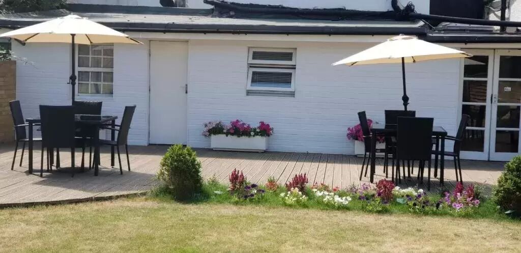 Un patio con dos mesas y sillas, un edificio blanco con ventanas y una puerta, y un macizo de flores variado.