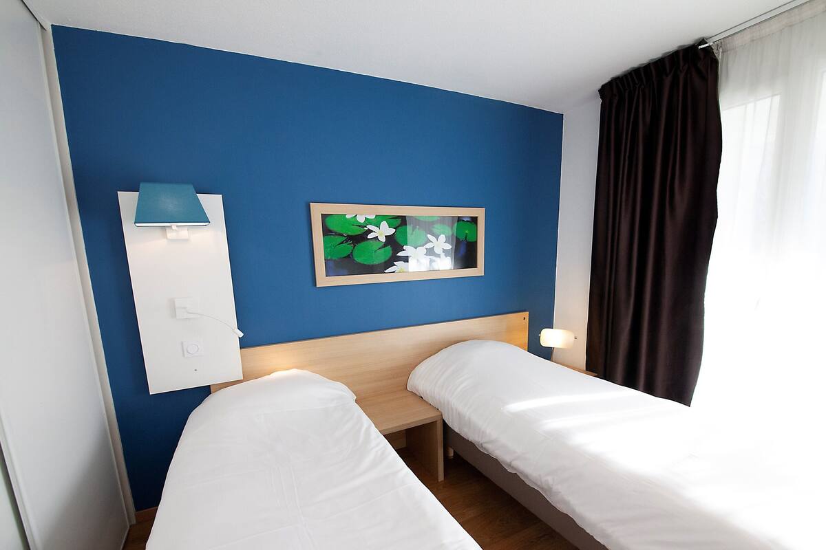 Habitación de hotel con dos camas individuales, una mesita de noche, un cuadro enmarco, una lámpara y una ventana con cortinas.
