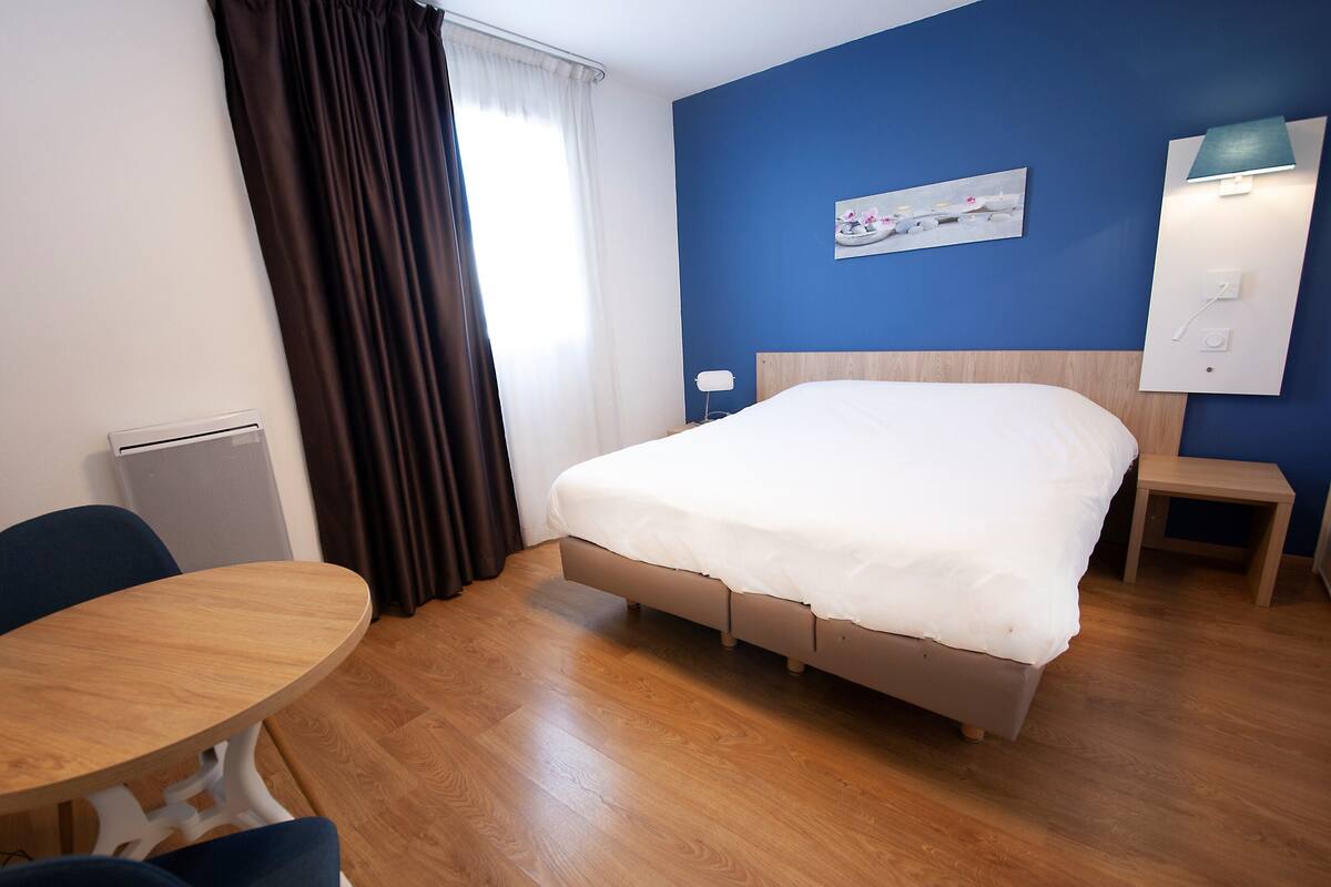 Habitación de hotel con cama, una mesa redonda, una silla, un radiador y una lámpara de pared.