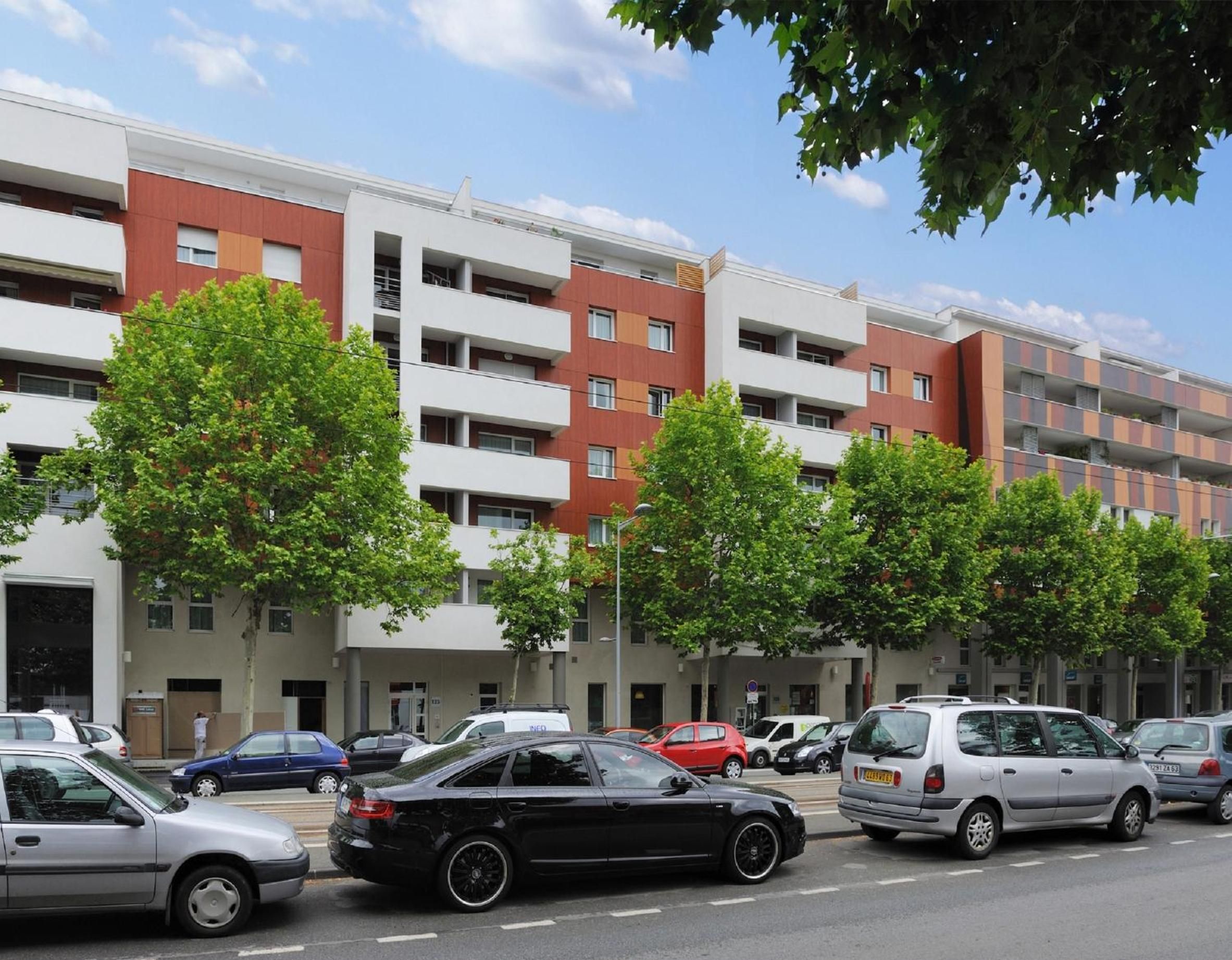 Un edificio residencial de varios pisos con balcones, coches aparcados y árboles.