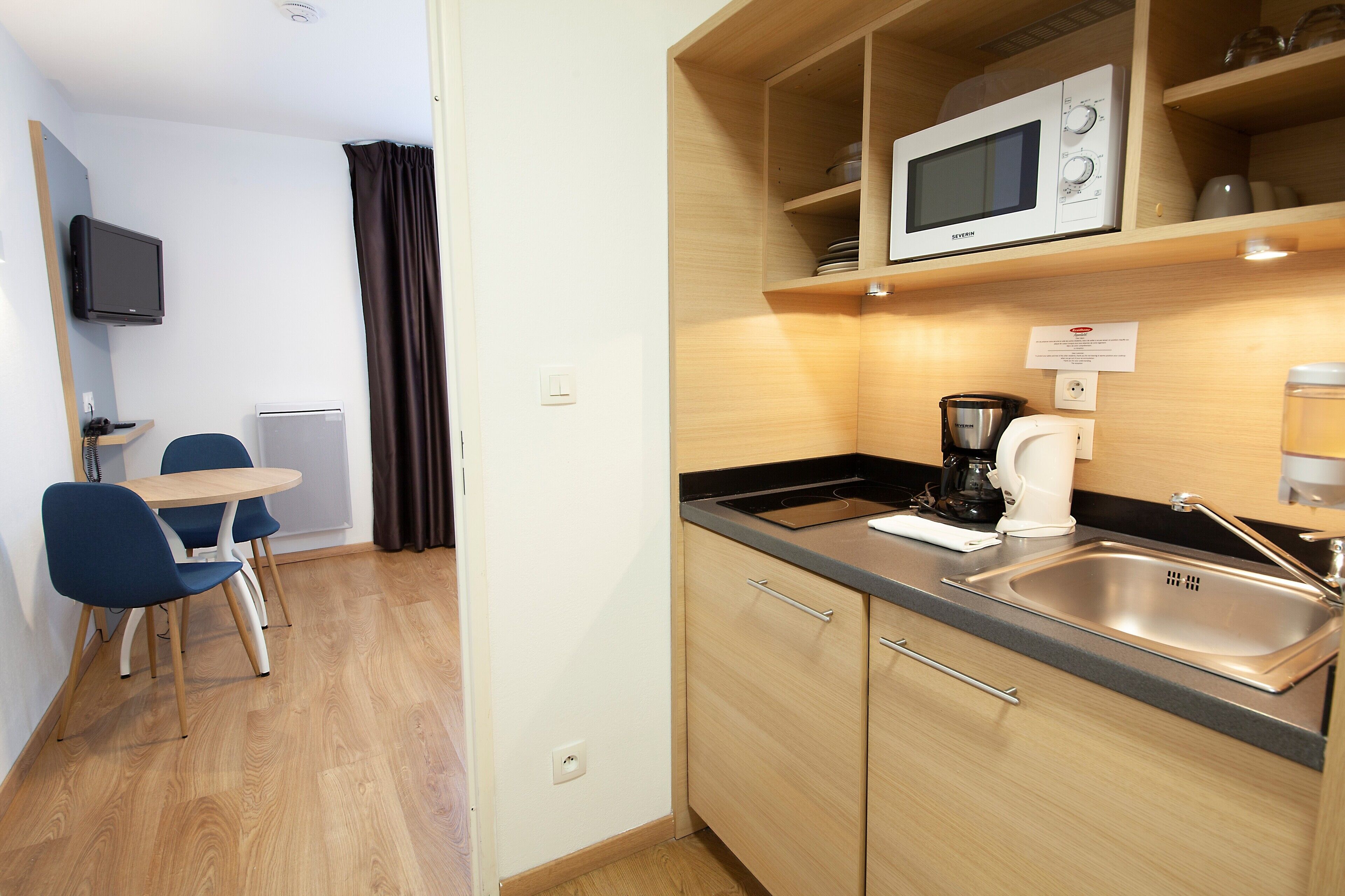 Una kitchenette compacta con microondas, cafetera y lavabo. Hay un pequeño comedor con mesa y sillas.