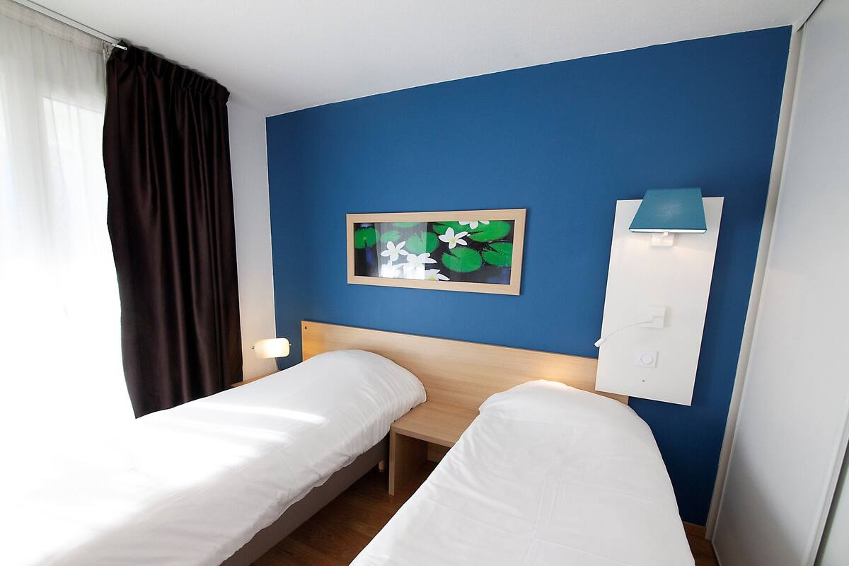 Habitación de hotel con dos camas individuales, una mesita de noche con lámpara, un enchufe en la pared y un cuadro con flores.