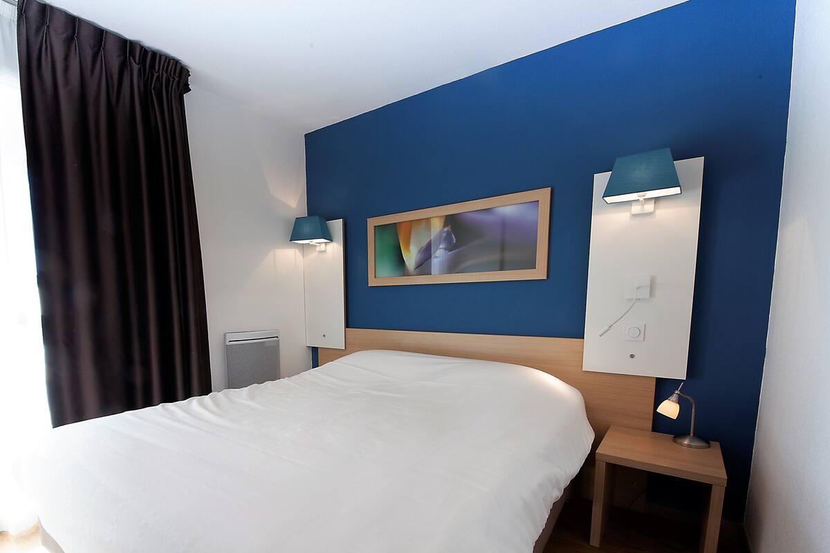Habitación de hotel con una cama grande, una mesita de noche con lámpara, un televisor montado en la pared y una ventana con cortinas.