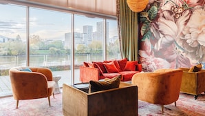 Uma sala de estar moderna com grandes janelas, vista para um rio e horizonte urbano, e um mural floral na parede.