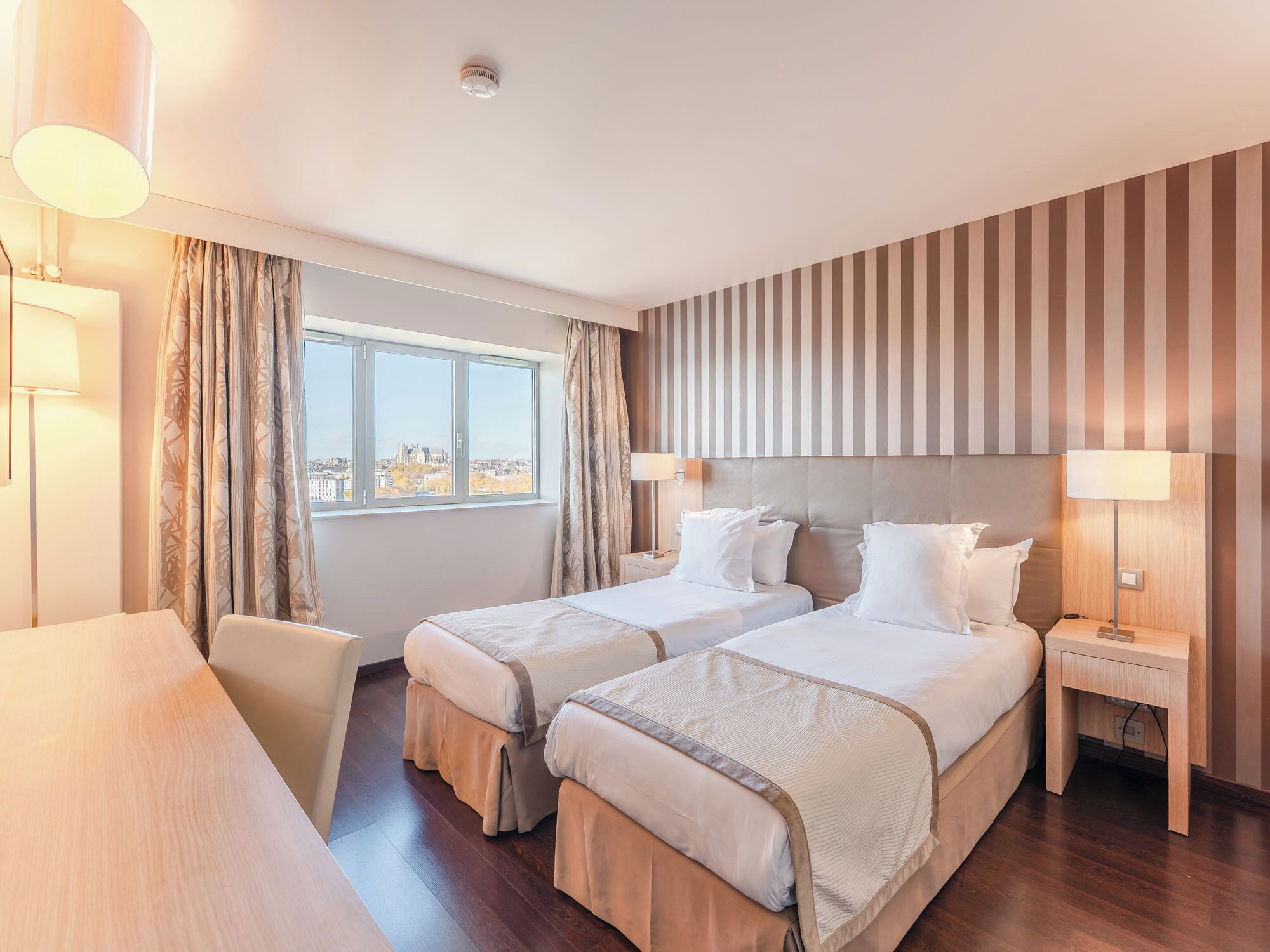 A hotel room with two beds, a desk, a chair, a window with a city view, and a striped wall.