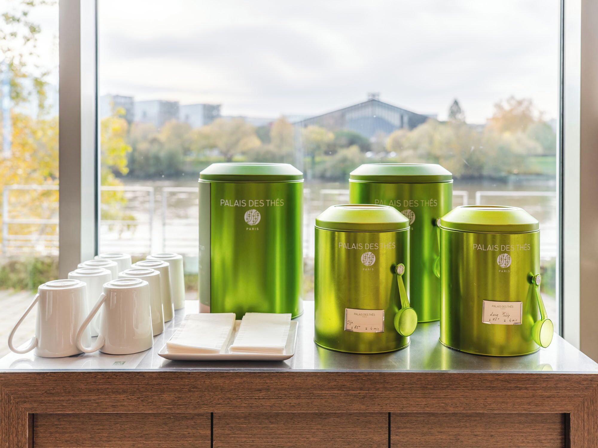 A kitchen counter with green thermal containers and white mugs, with a view of trees and buildings outside.
