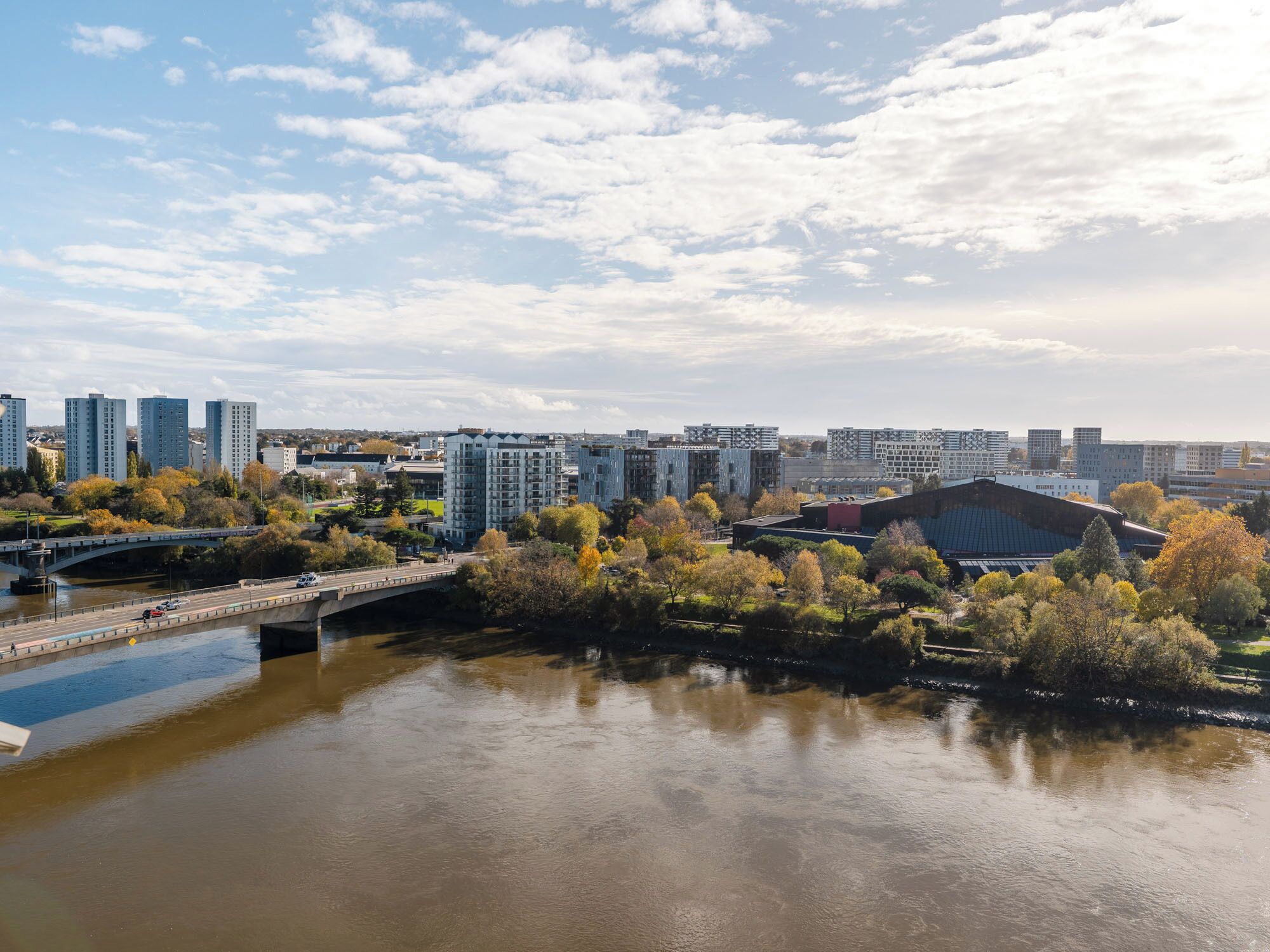 A riverside cityscape with modern buildings, a bridge, and autumn foliage.