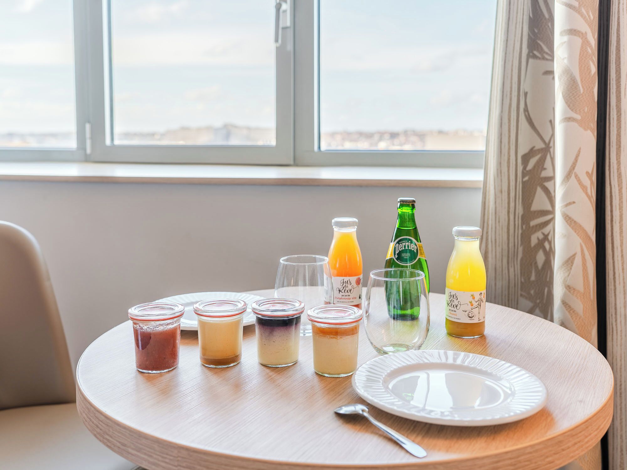 Uma mesa de madeira redonda com várias bebidas e um prato, colocada perto de uma janela com vista para a praia.