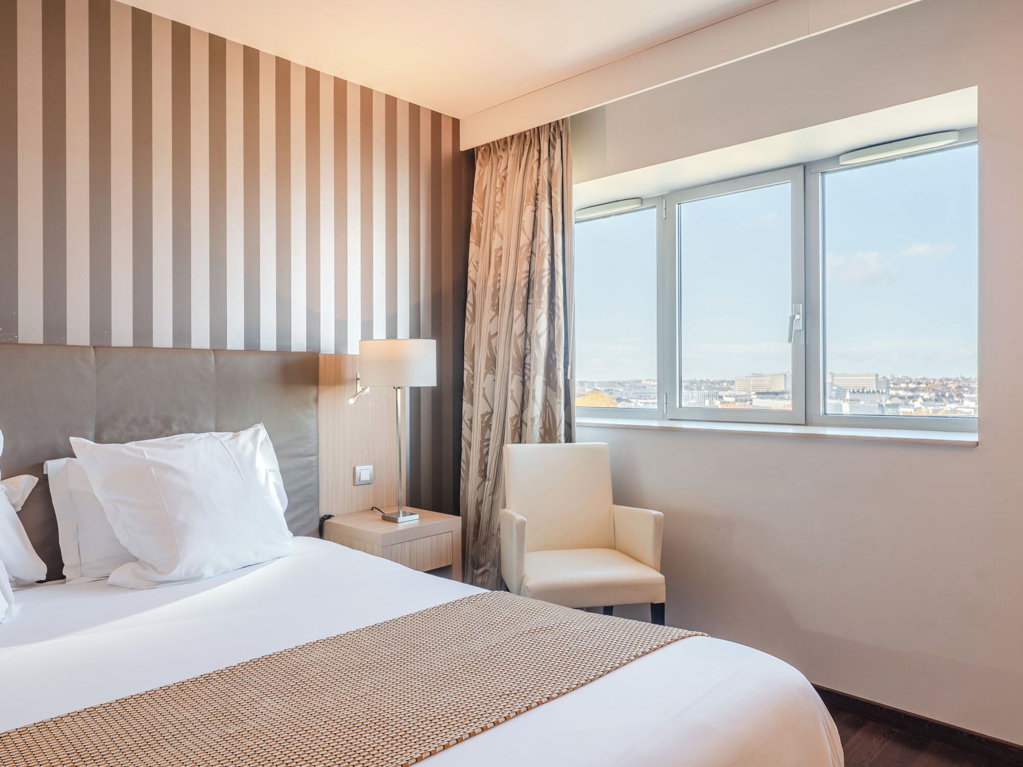 Camera d'albergo con un letto grande, una sedia, un comodino, una lampada e una finestra con vista sulla città.