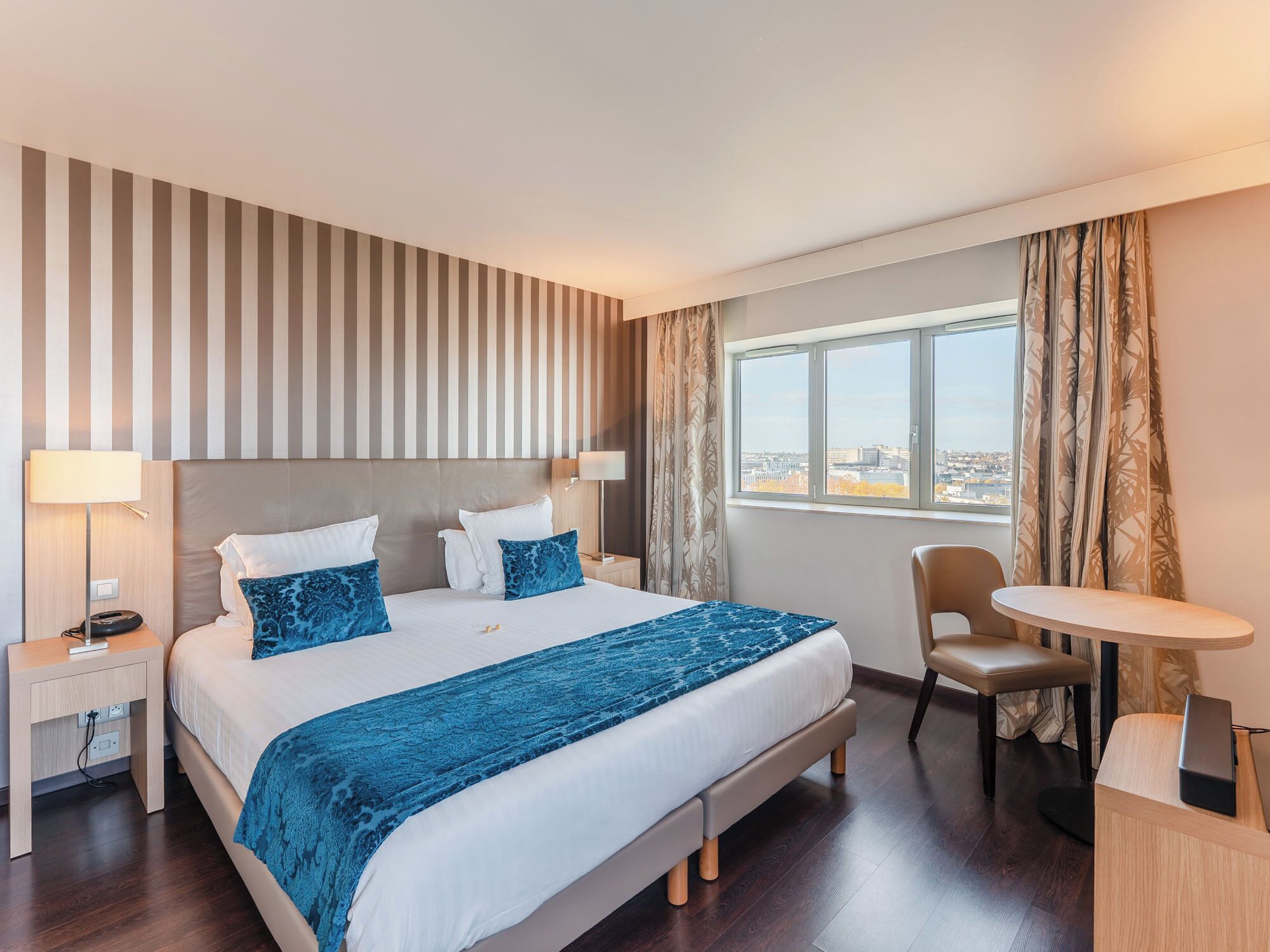A hotel room with a large bed, a desk, a chair, and a view of the city through the window.