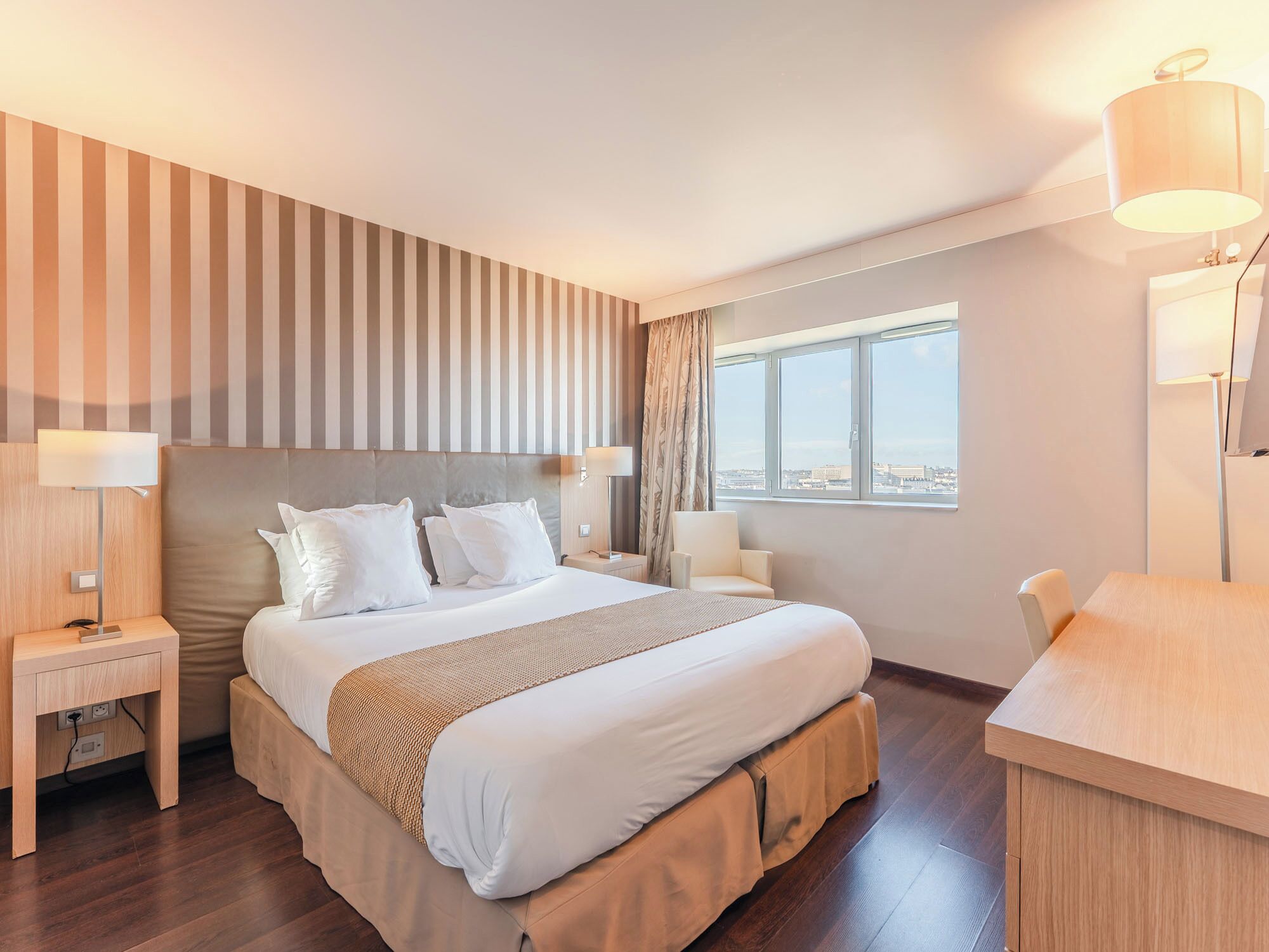 A hotel room with a large bed, two bedside tables, a desk, and a window with a city view.