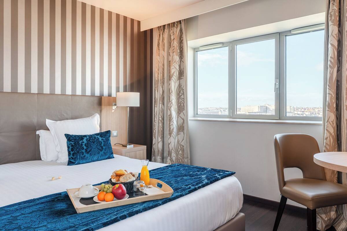 Une chambre d’hôtel avec un lit, un plateau de petit-déjeuner avec des fruits et du jus, une chaise et une vue sur la ville par la fenêtre.