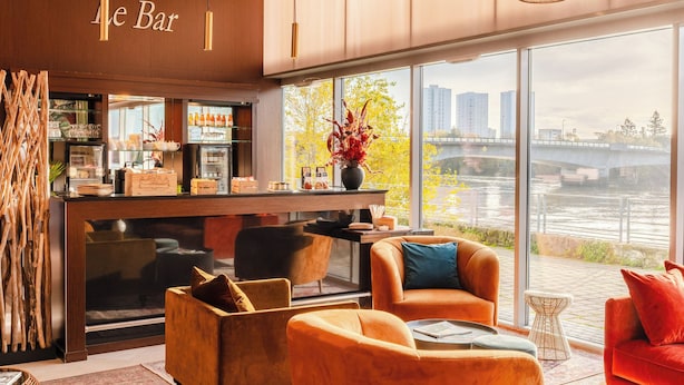 Uma área de bar moderna com vista para um rio e uma ponte, mobiliada com sofás laranja e uma máquina de café.