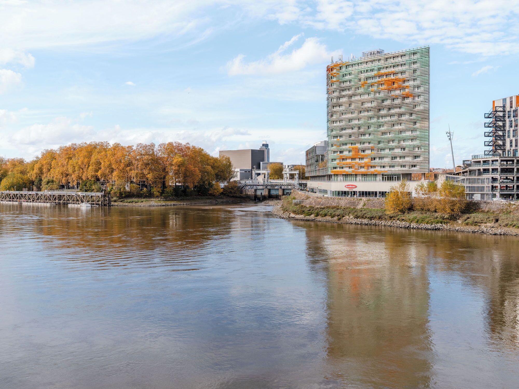 A modern building with a glass facade, located beside a river with autumn trees in the background.