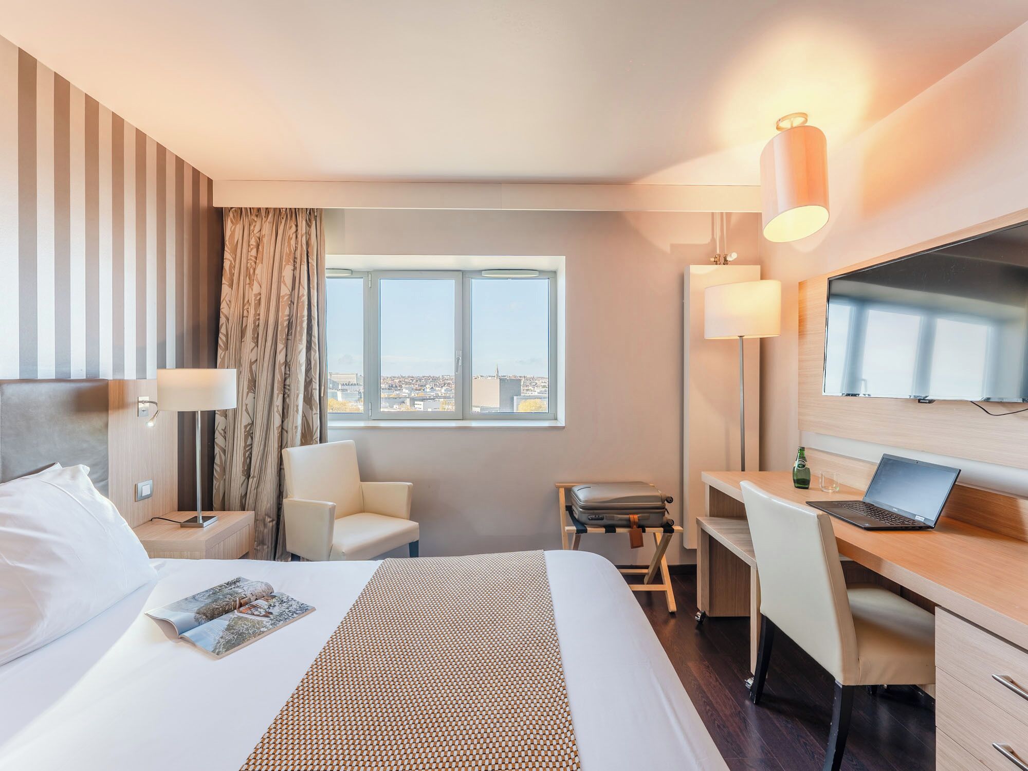 A hotel room with a bed, a desk, a chair, a TV, and a window with a city view.