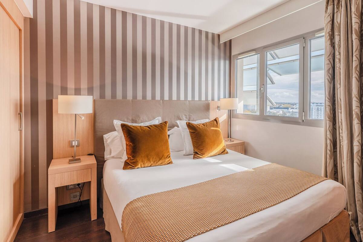 Une chambre d’hôtel avec un grand lit, deux tables de chevet, une fenêtre avec des rideaux et un mur à rayures.