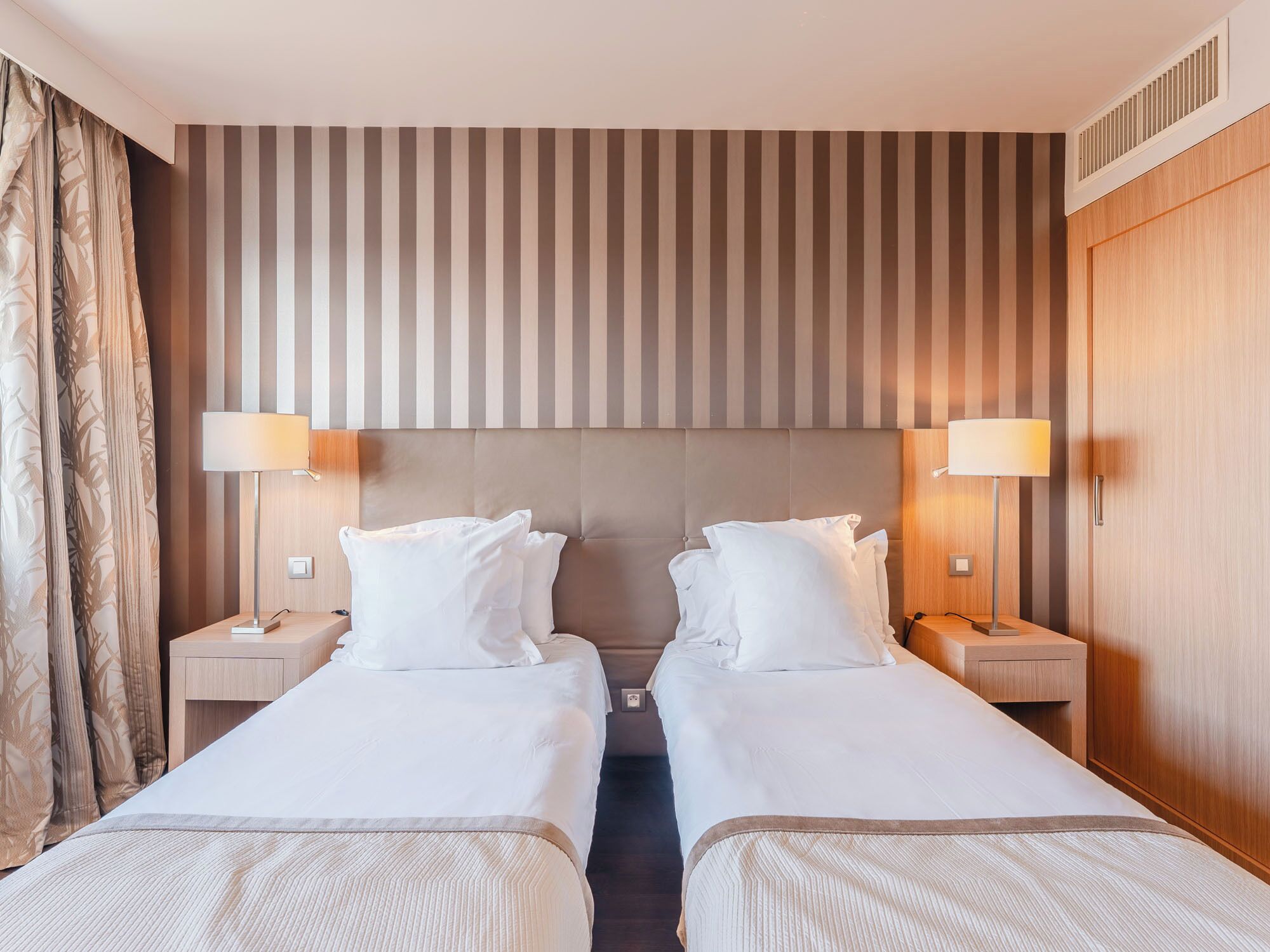 Habitación de hotel con dos camas, cabecera a rayas, lámparas de noche y un armario de madera.