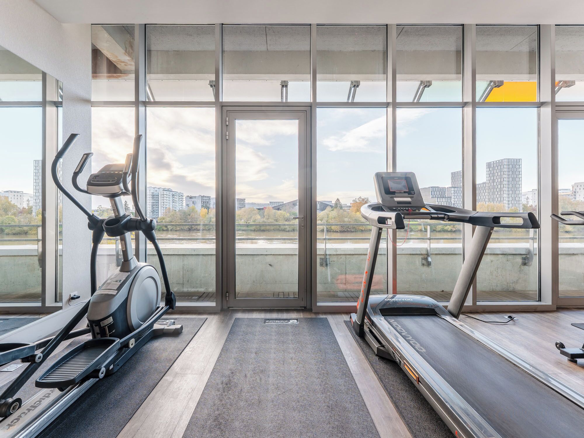 A gym with treadmills and an elliptical machine, large windows with a city view, and a concrete floor.