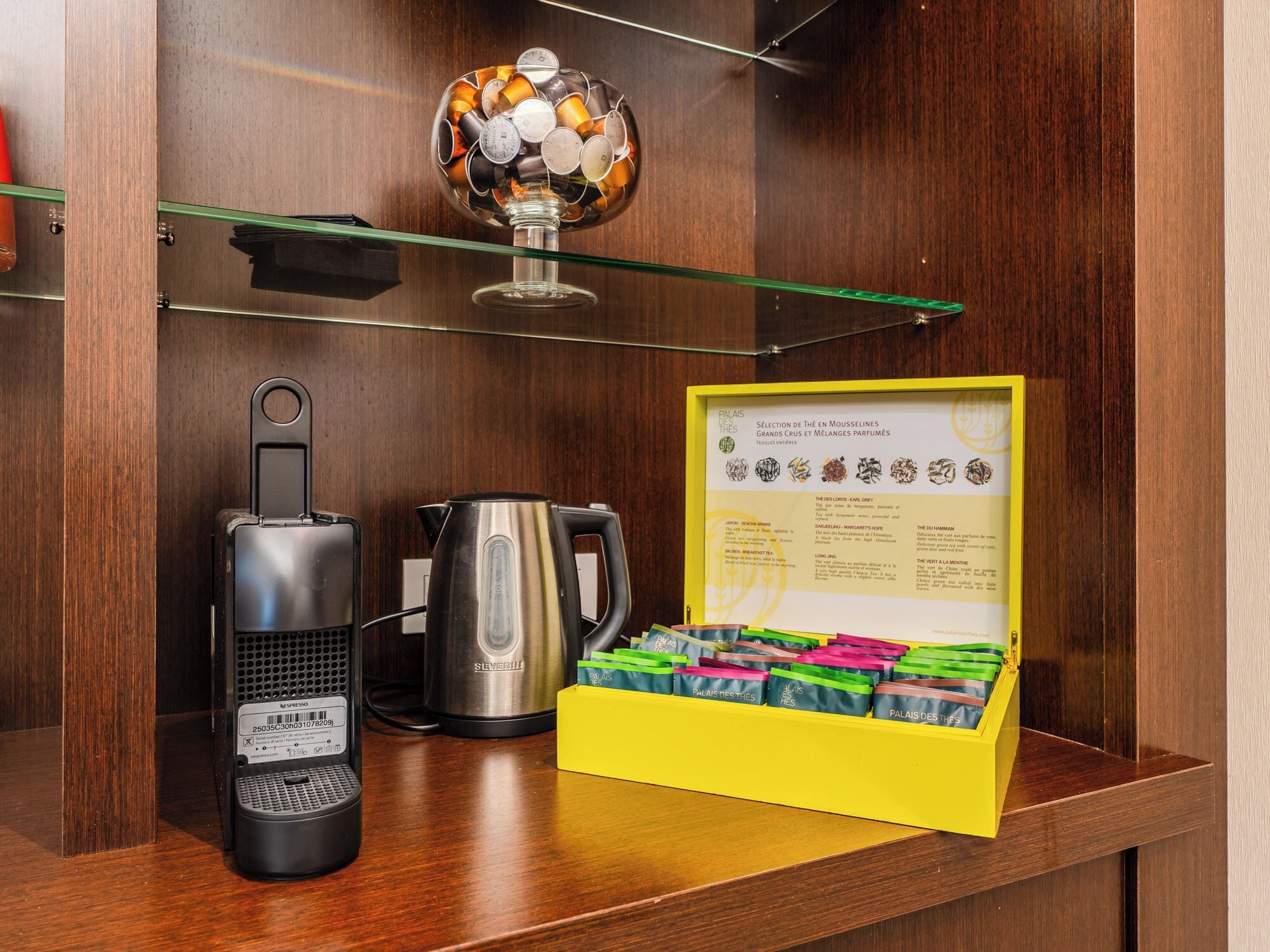 A coffee maker, a kettle, and a selection of tea boxes on a wooden shelf.