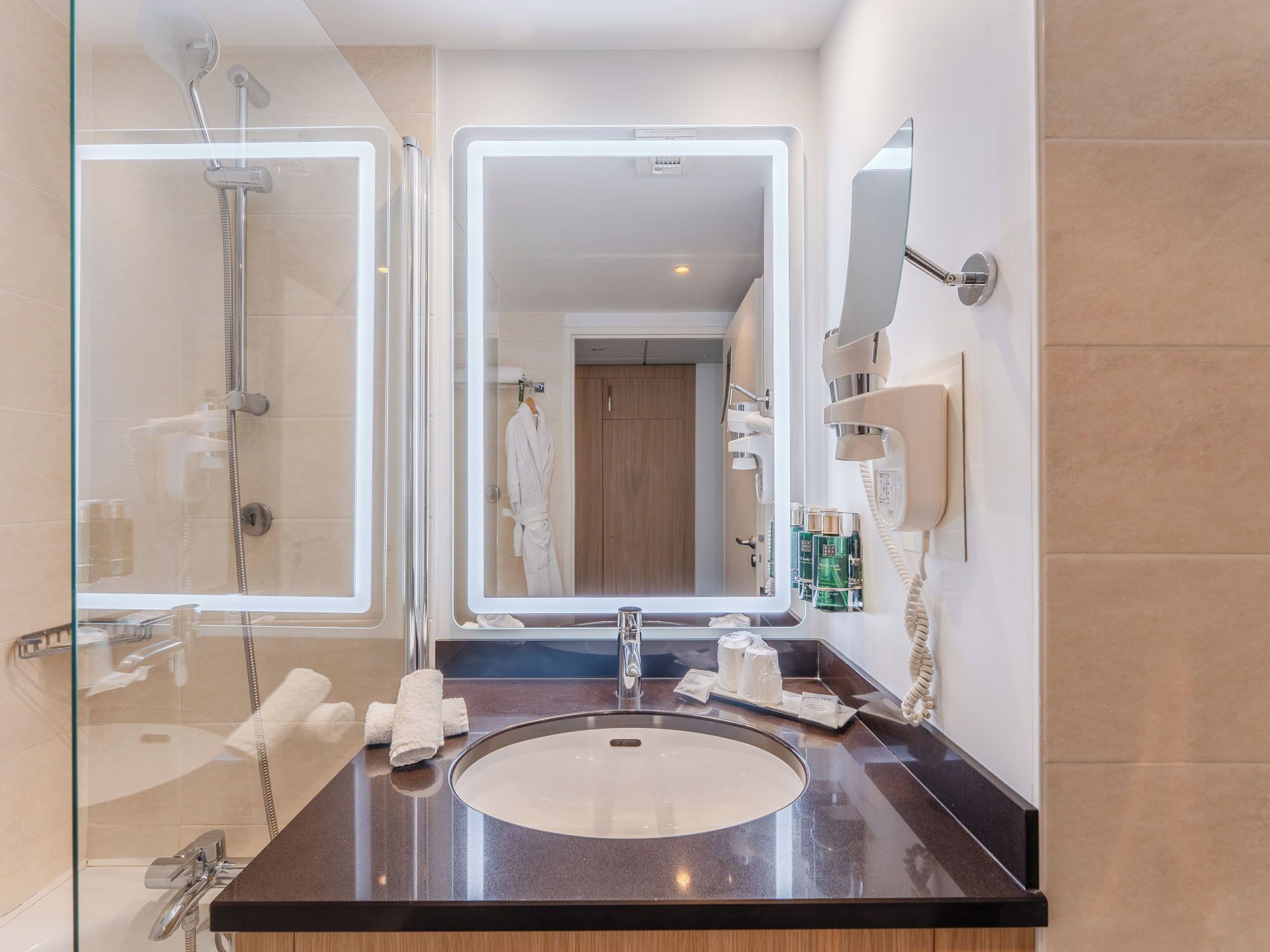 Een moderne badkamer met een douchecabine van glas, een grote spiegel, een wastafel met toiletartikelen, en een haardroger aan de muur.