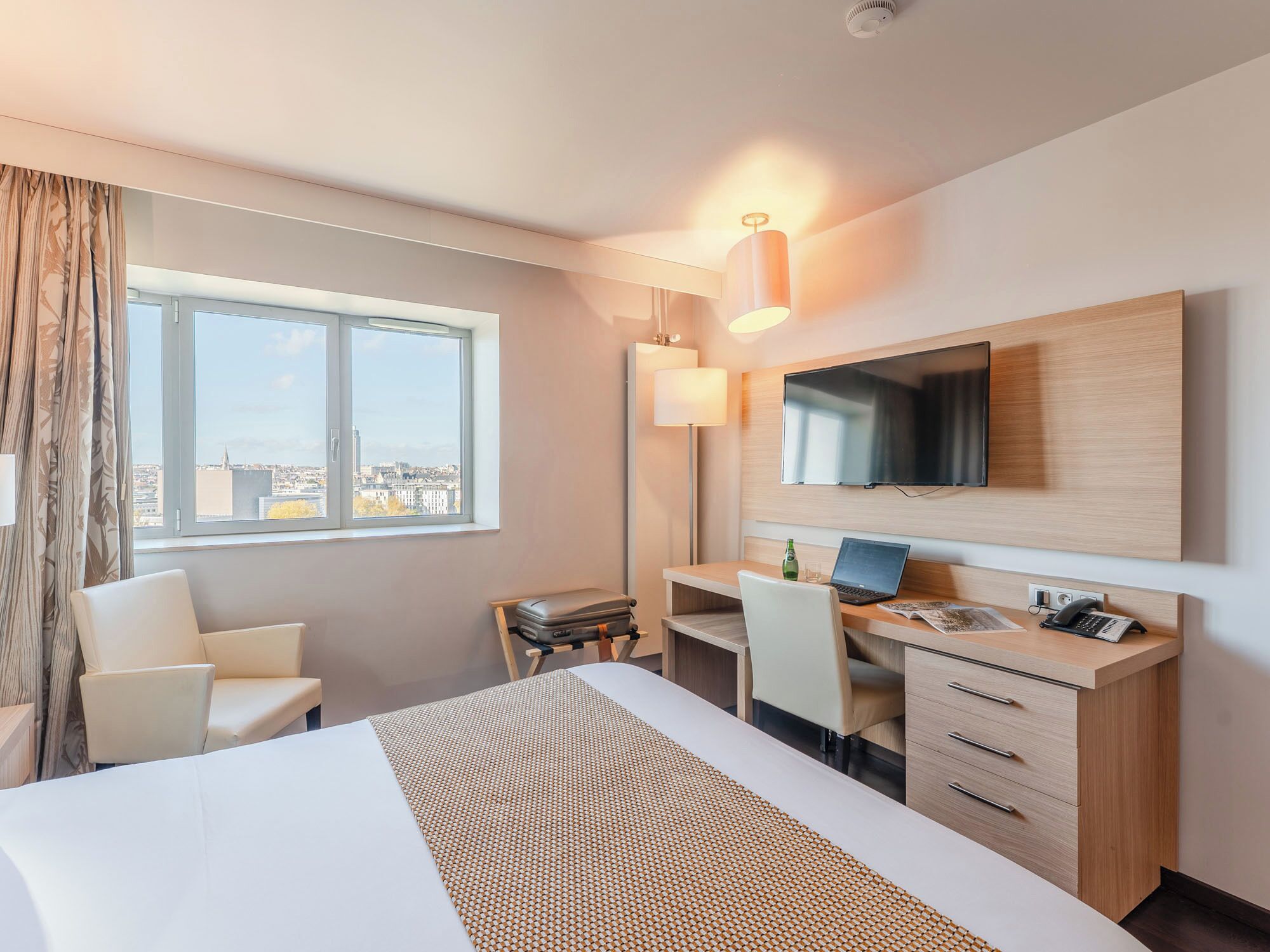 A modern hotel room with a large bed, a desk with a laptop, a chair, a flat-screen TV, and a city view.