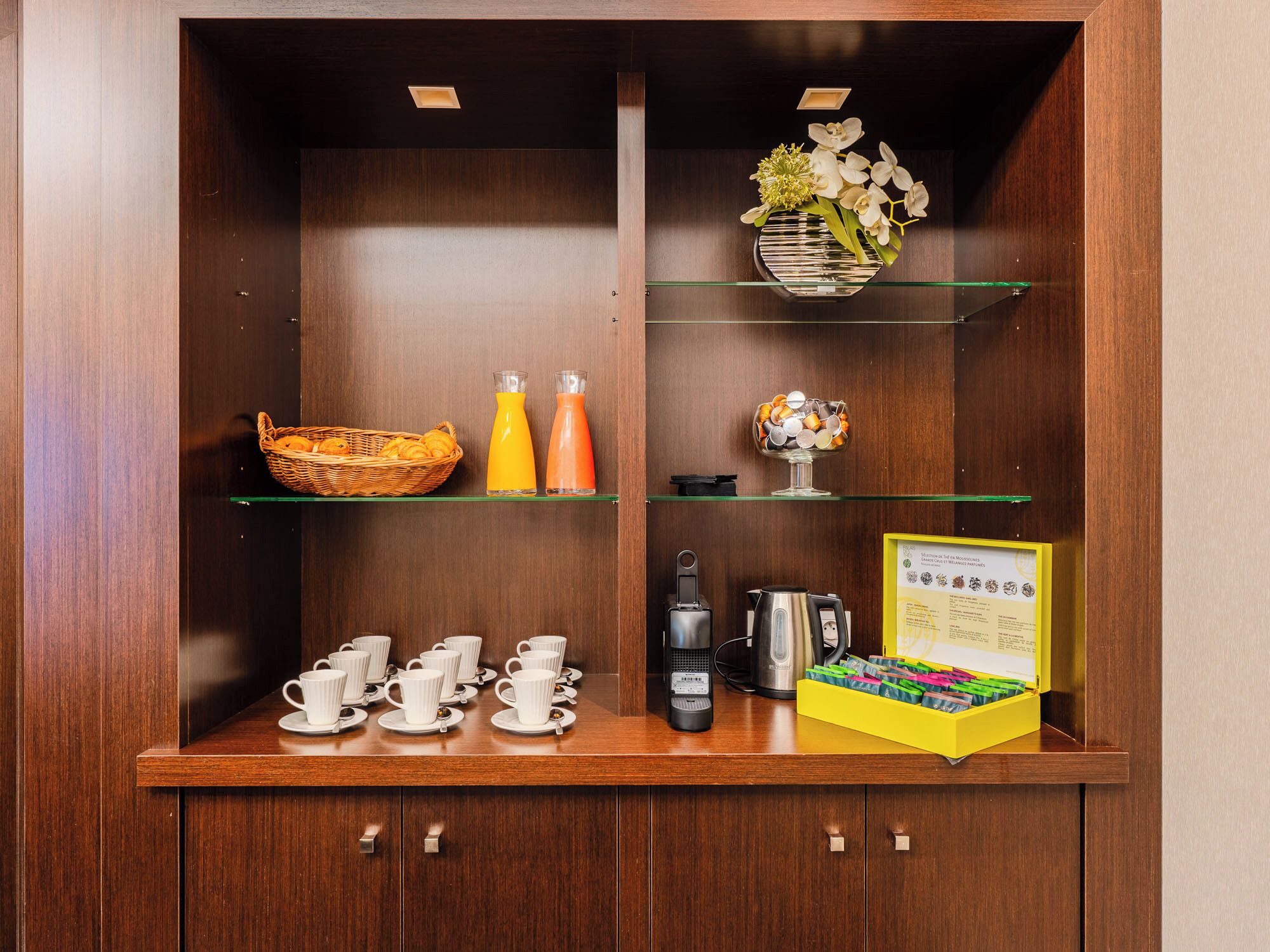 Un meuble en bois doté d’étagères en verre, contenant un panier d’oranges, deux bouteilles de jus d’orange, une machine à café, une bouilloire, une boîte de stylos de couleurs vives et un certificat encadré.
