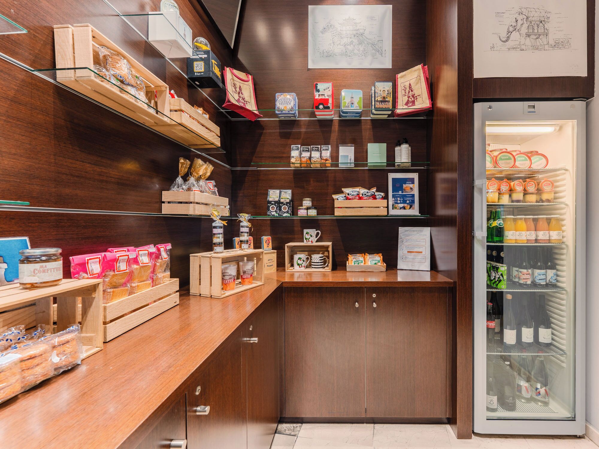 Un bar de aperitivos compacto y ordenado, con estantes de madera que exhiben diversos productos envasados, un mostrador refrigerado con bebidas y una pequeña zona de estar con barra.