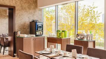 Un espace repas moderne équipé d’une machine à café, de distributeurs de boissons vertes et offrant une vue sur les arbres aux couleurs automnales grâce à de grandes fenêtres.