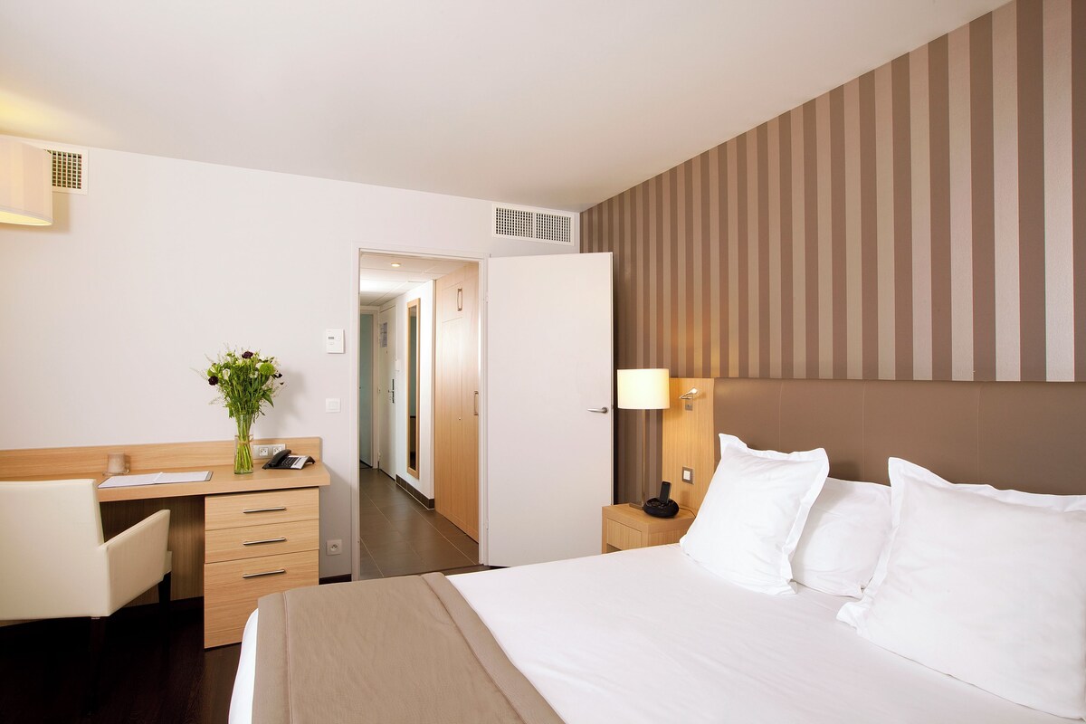 A hotel room with a bed, a desk, a chair, a nightstand, and a lamp.