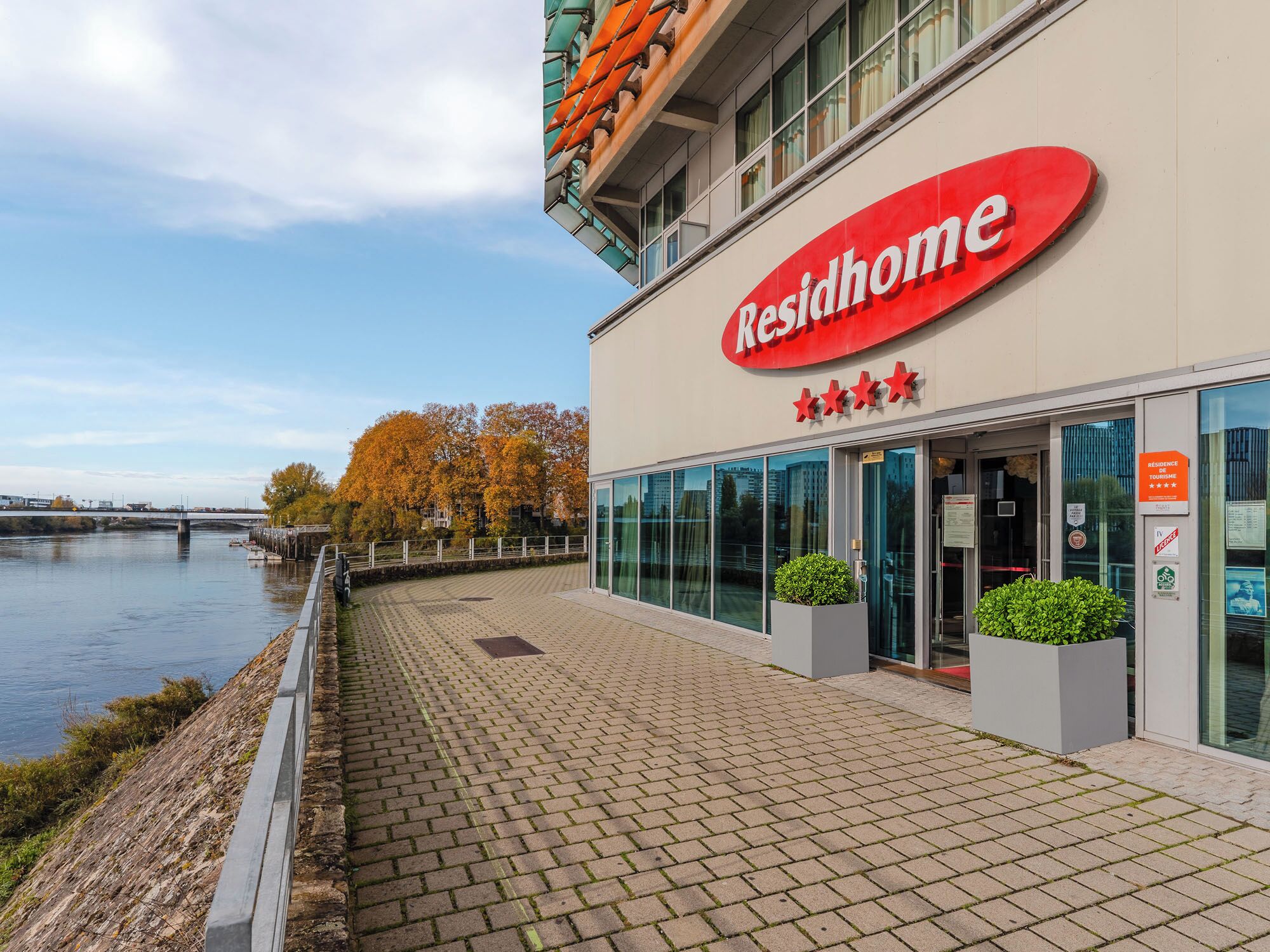 Un edificio moderno con un letrero que dice "Residhome", con fachada de vidrio y un paseo pavimentado junto a un río.