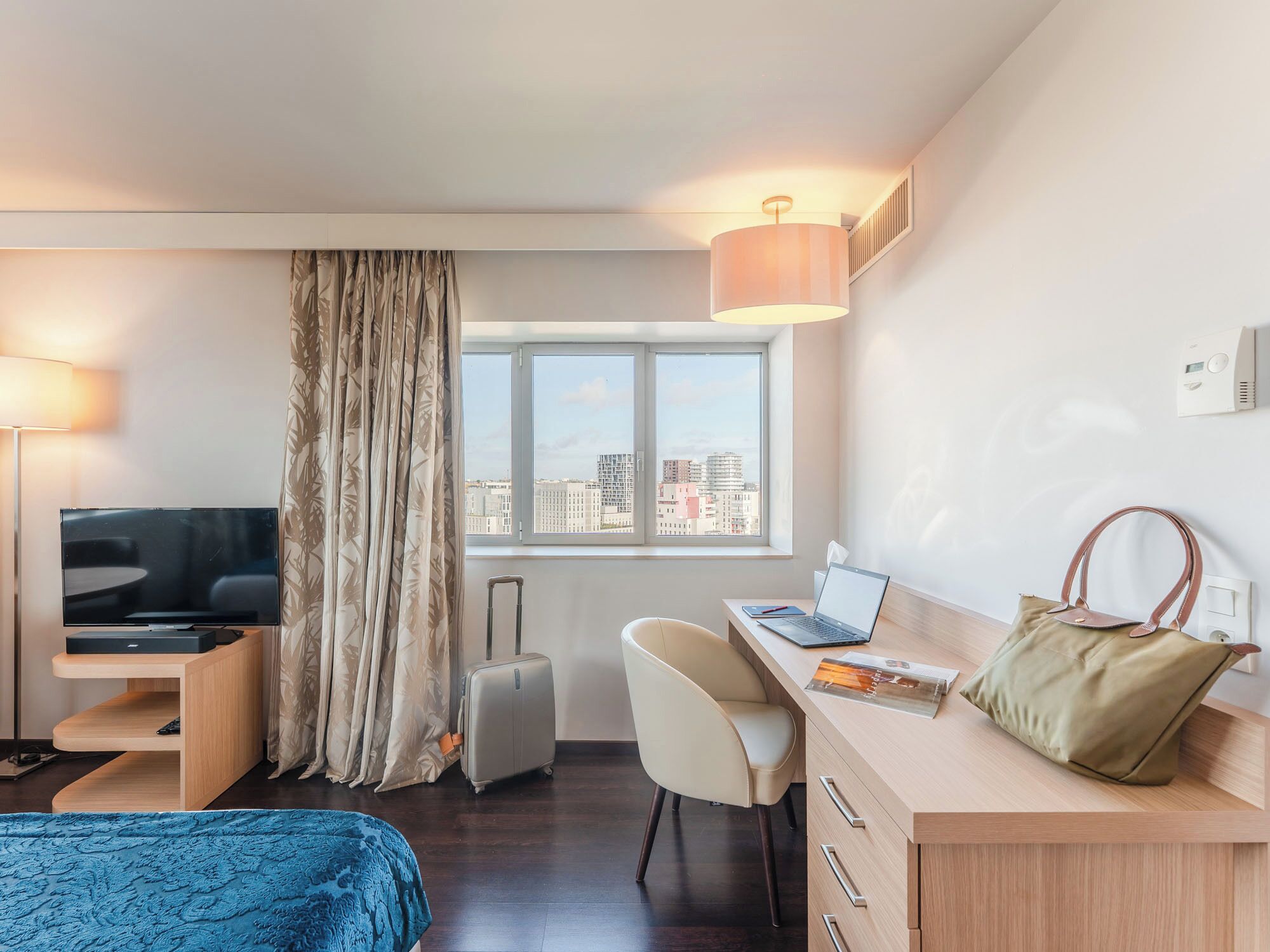 A hotel room with a bed, a desk with a laptop, a chair, a TV, a suitcase, a lamp, a window with a view of buildings, and a handbag.