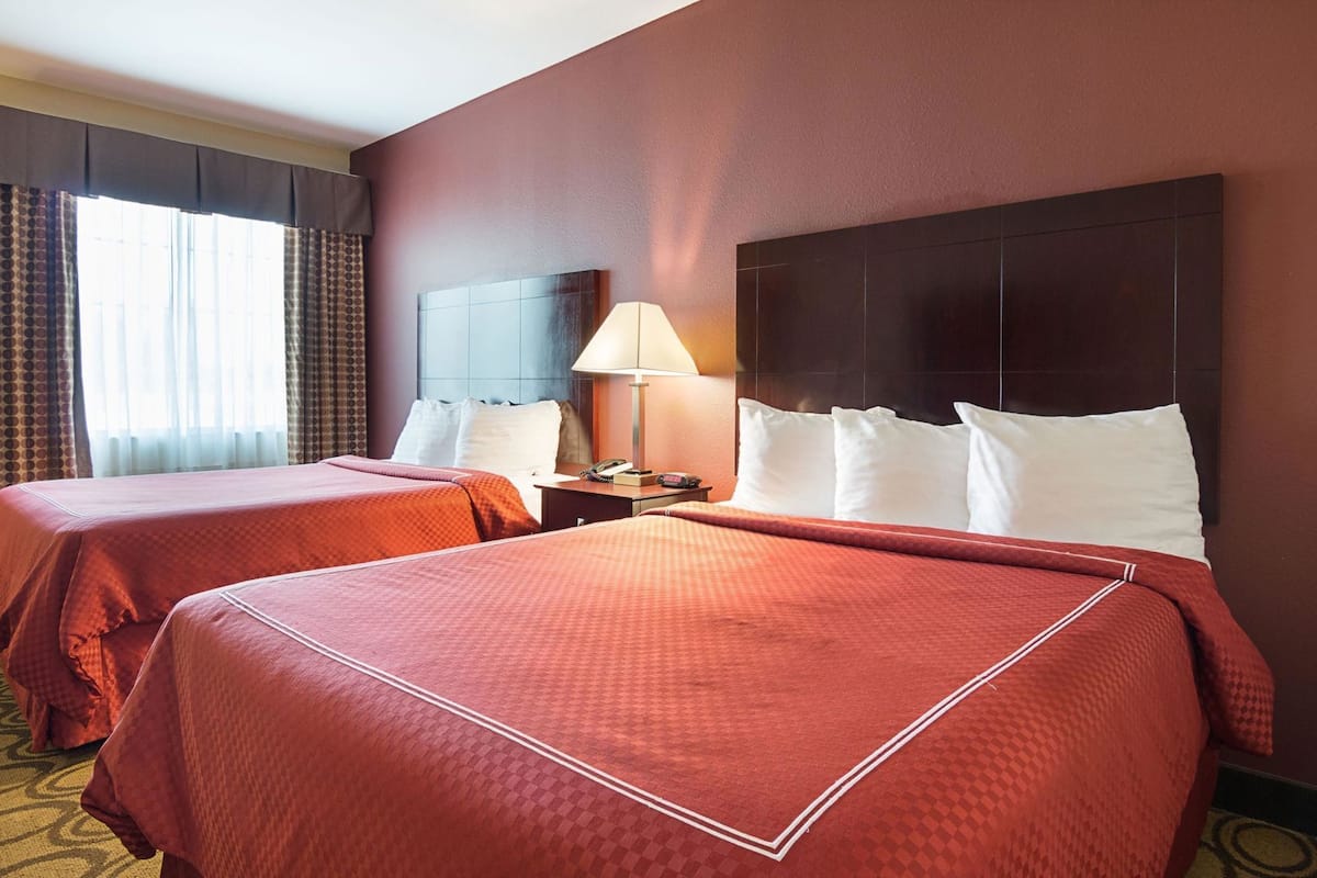 A hotel room with two beds, a nightstand, a lamp, and a large window with curtains.