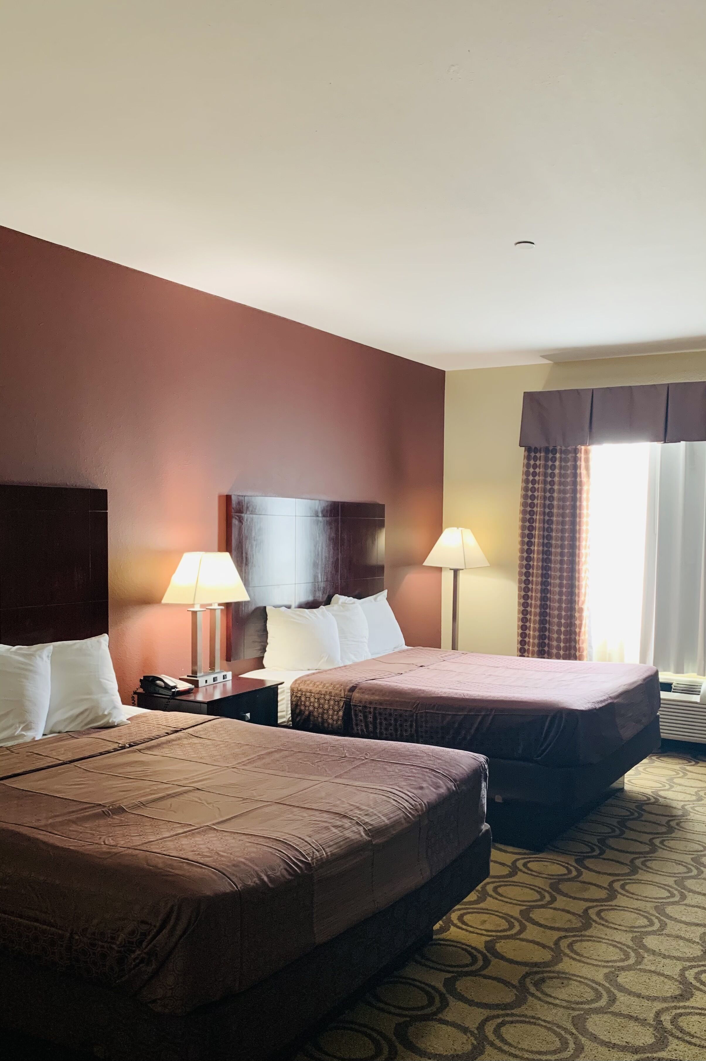 A hotel room with two beds, a nightstand with a lamp, and a window with curtains.
