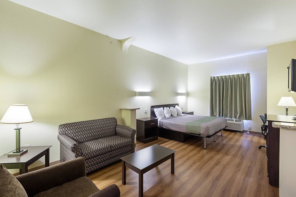 A hotel room with a bed, a sofa, a small table, and a desk. There are two lamps on the nightstands and a window with curtains.